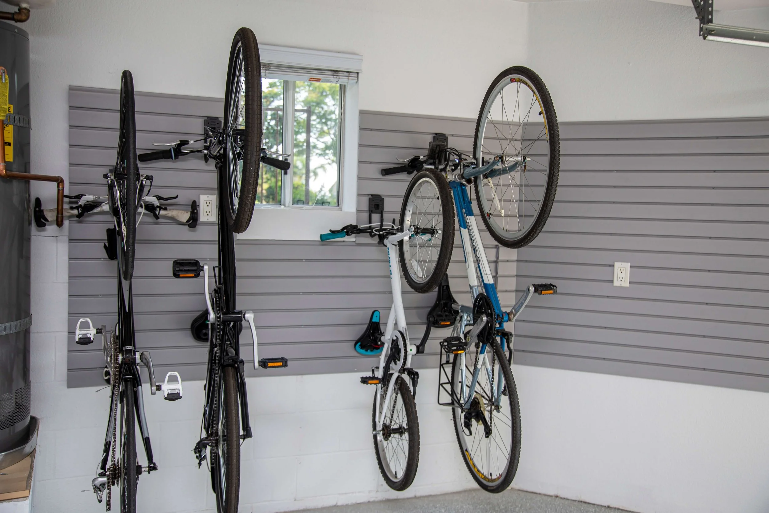 Several bicycles mounted on a wall with gray slat wall panels in a garage, with a window showing greenery outside.