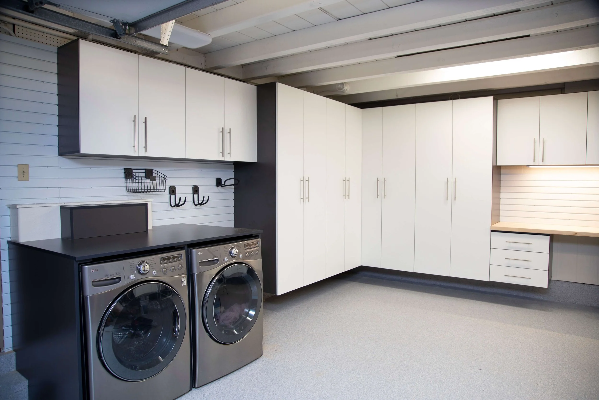 A laundry room with white cabinets, a countertop, and two front-loading washing machines and dryers. The room has a sloped ceiling with visible beams and a white shiplap wall.