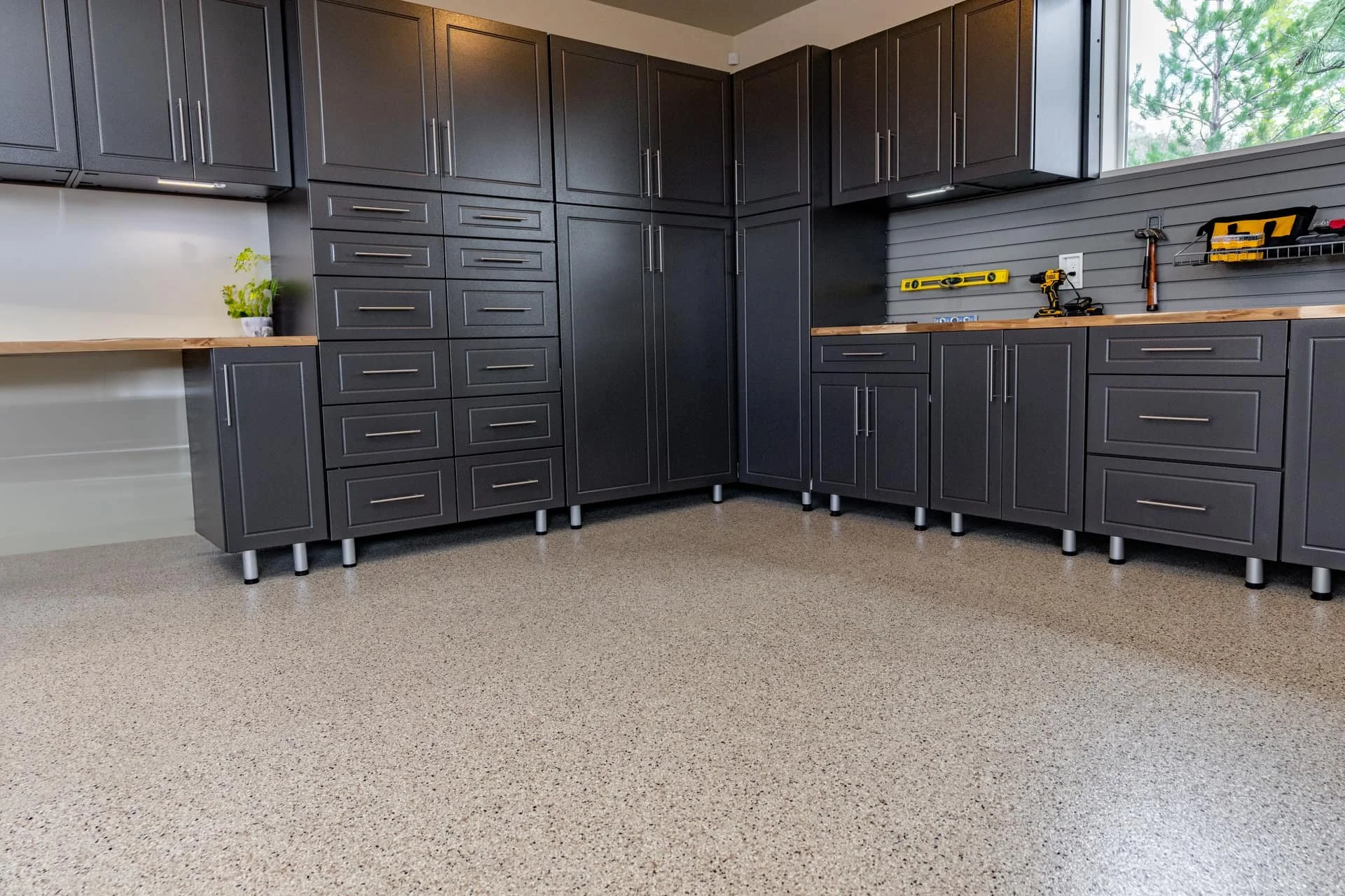 Kitchen with dark gray cabinets, a wooden countertop, and tools hanging on the wall. A plant in a white pot sits on the countertop near the left side. There is a large window on the right providing natural light, and the floor has a speckled, beige surface.