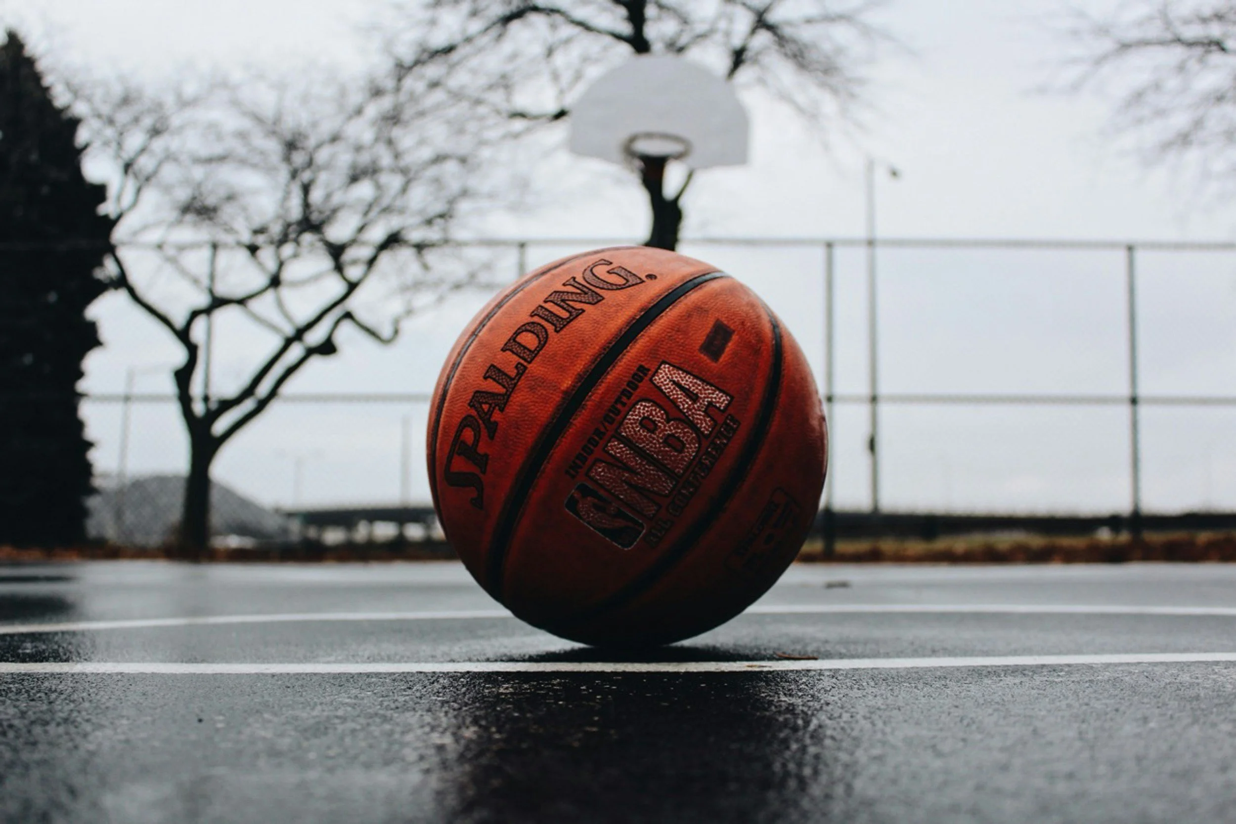 A basketball on an empty outdoor court with trees and a basketball hoop in the background on a cloudy day.