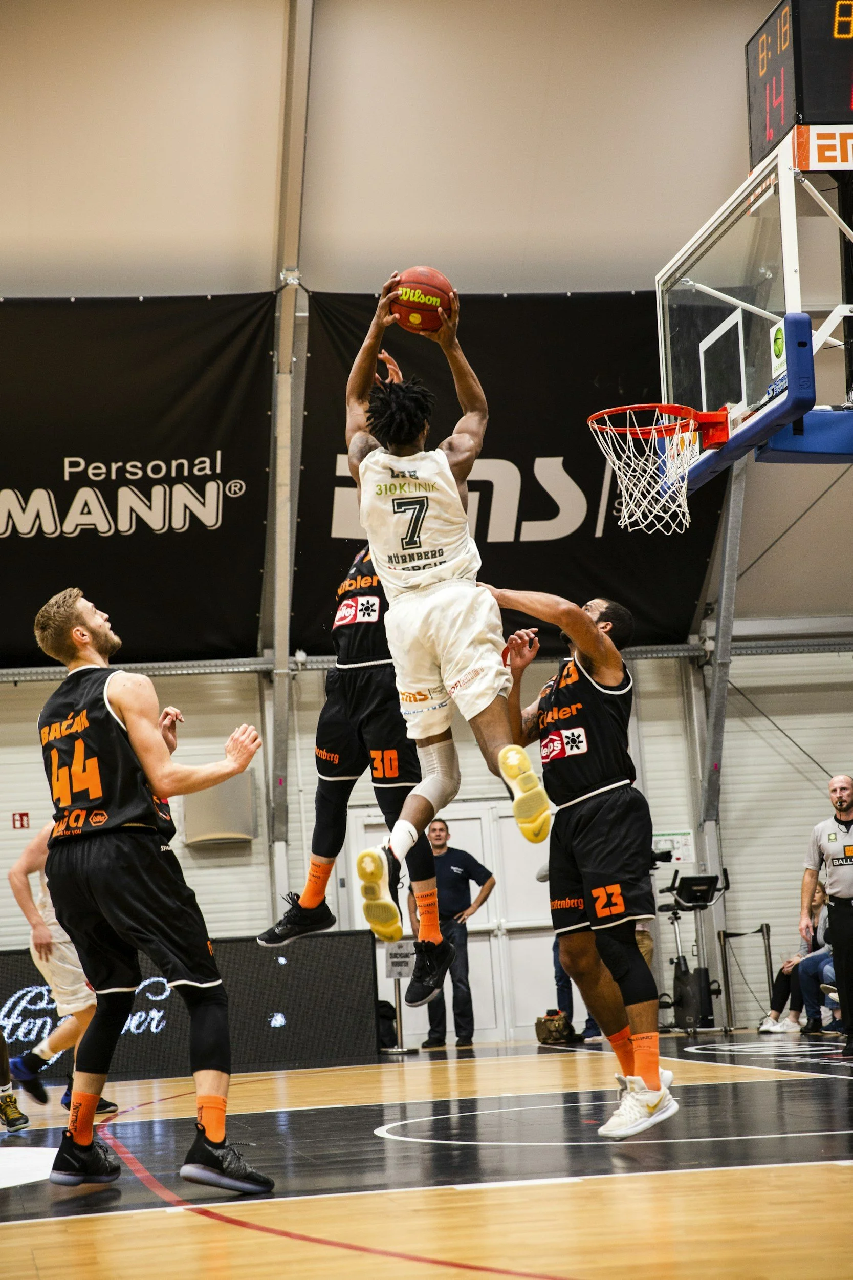 A basketball player in a white uniform with the number 7 is jumping to dunk the ball while being defended by three players in black uniforms on an indoor court.