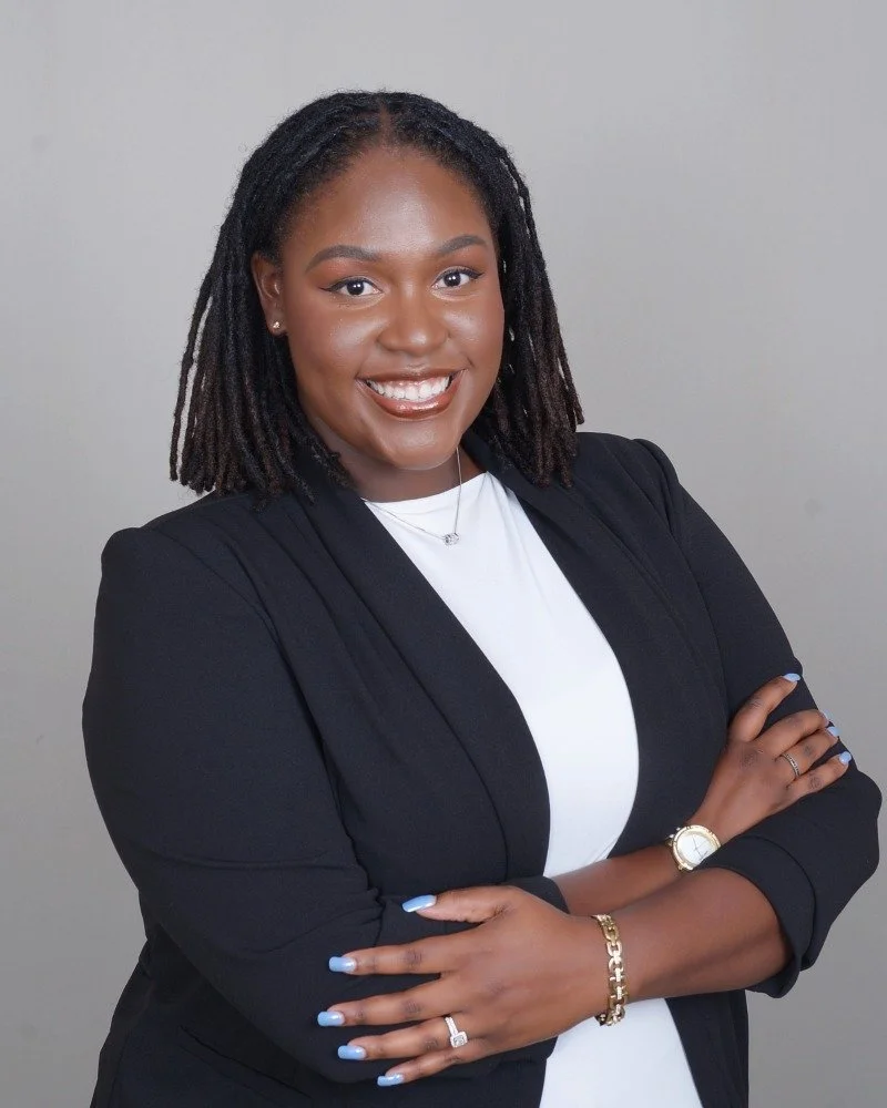 Professional woman with braided hair, wearing a black blazer and white top, smiling, standing against a neutral background