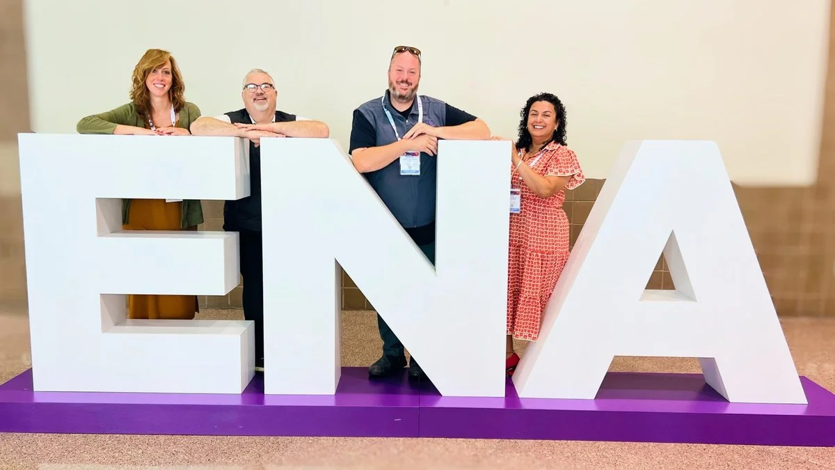 Four people smiling and leaning on large white letters spelling "ENNA" with a purple base, in an indoor setting.