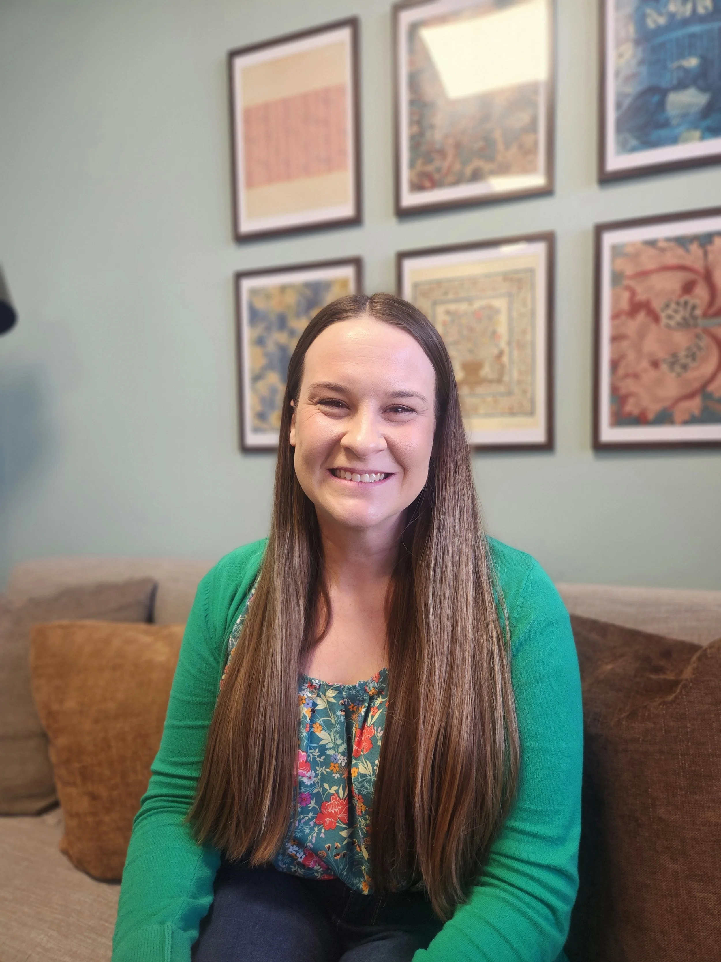 A woman with long brown hair smiling, sitting on a beige sofa with brown pillows in a room with framed artwork on the wall behind her.