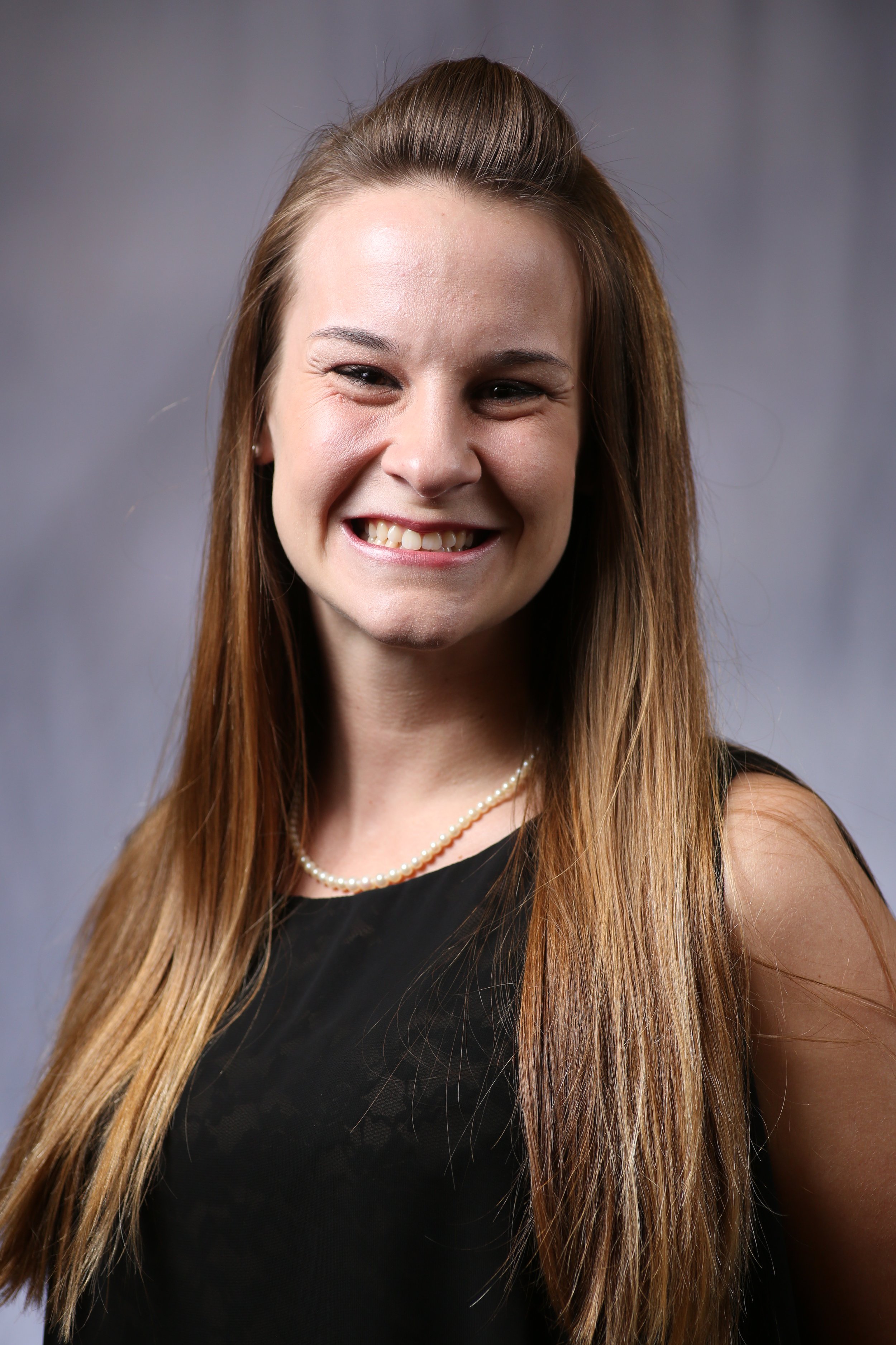 A woman with long, light brown hair styled with a small section pinned back, wearing a black dress with a lace pattern and a pearl necklace, smiling against a gray background.
