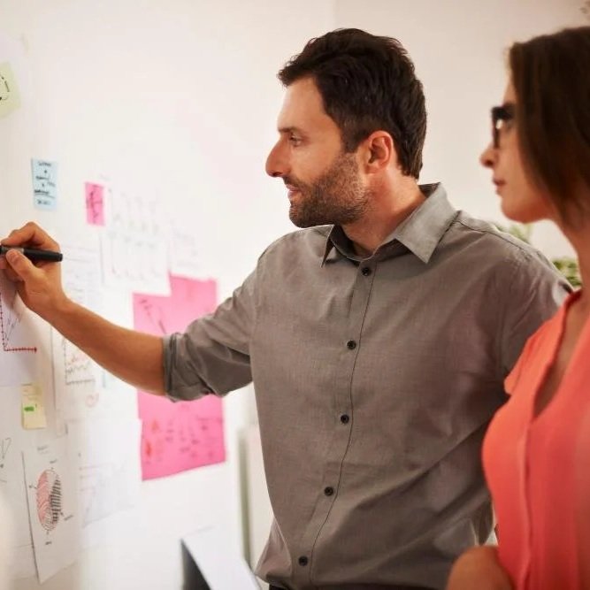 A man with dark hair and a beard writing on a whiteboard in a meeting room, with a woman with glasses and brown hair standing next to him, both engaged in a discussion.