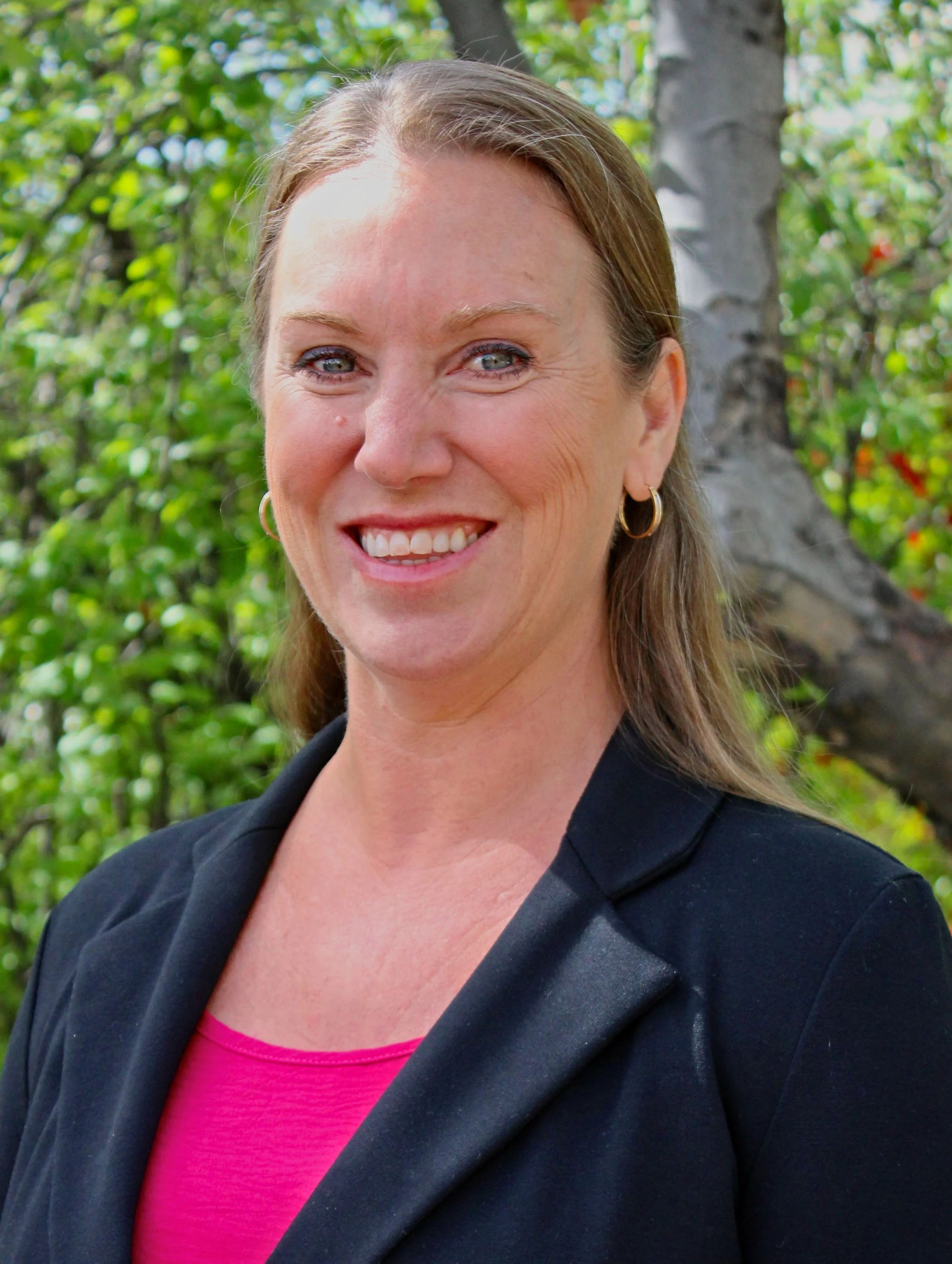 Portrait of a smiling woman with brown hair, wearing gold hoop earrings, black blazer, and pink top, standing outdoors with green foliage and tree in the background.