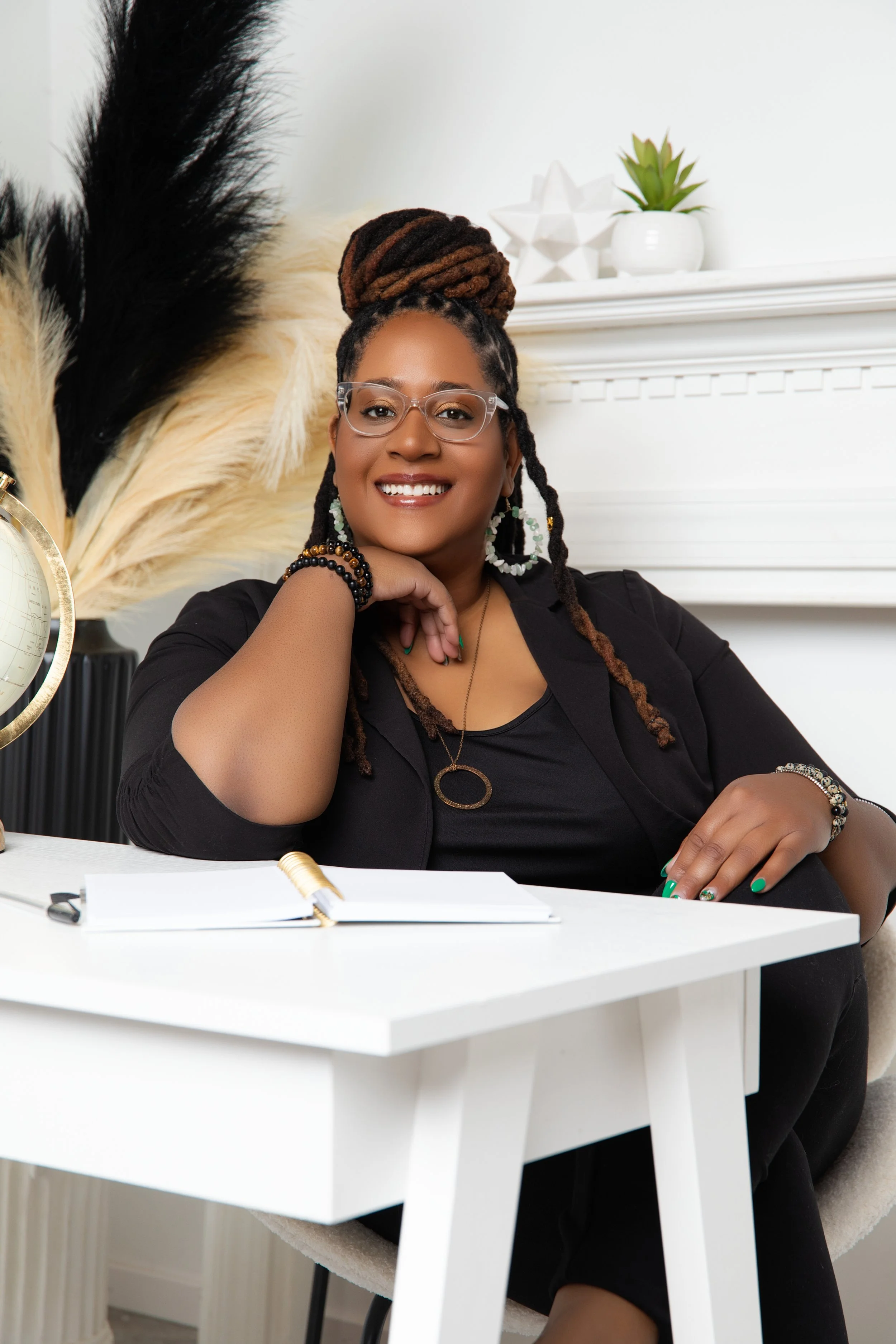 A smiling woman with glasses and dreadlocks in a bun, wearing black clothing and jewelry, sitting at a white desk with a notebook and pen, in a cozy office.