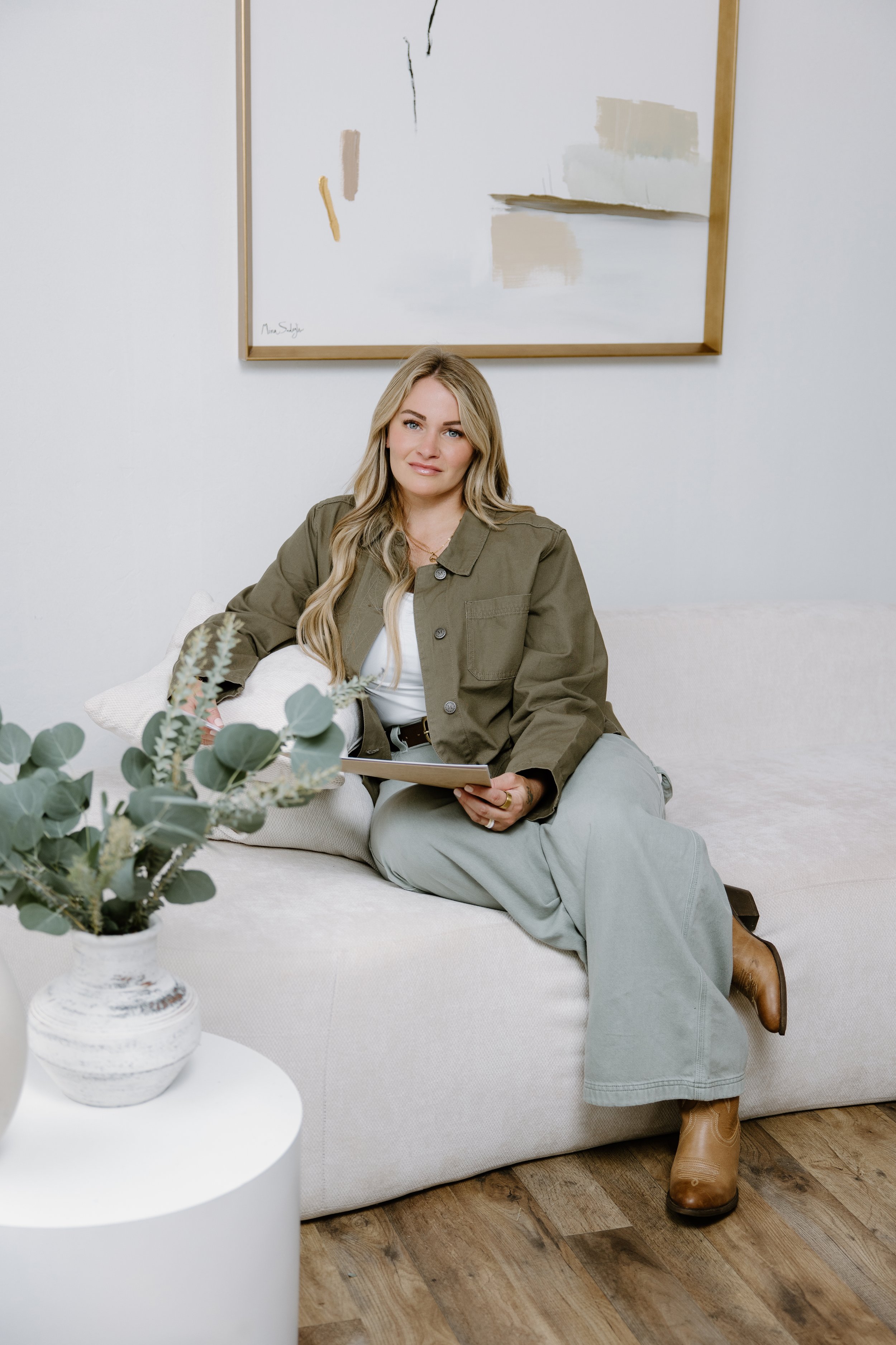 A woman sitting on a white sofa with a light-colored pillow, holding a closed laptop, in a room with a white wall, abstract painting above her, a small white table with a potted plant in the foreground, and hardwood flooring. She wears a beige jacket, gray pants, and tan boots.