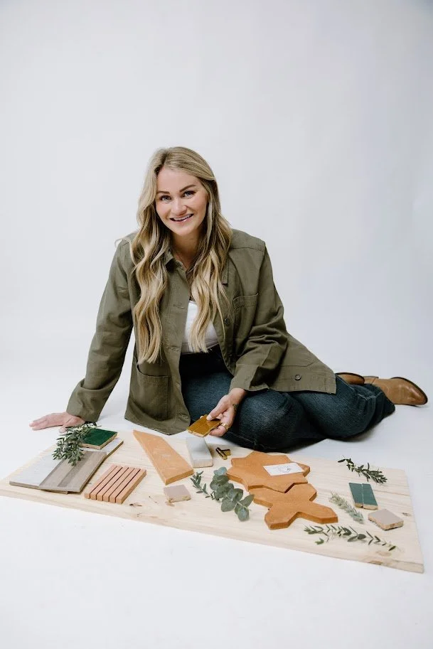 A woman with long blonde hair, wearing a green jacket, white top, and jeans, is sitting on the floor next to a large wooden board with various tile samples and greenery for a home renovation or interior design project.