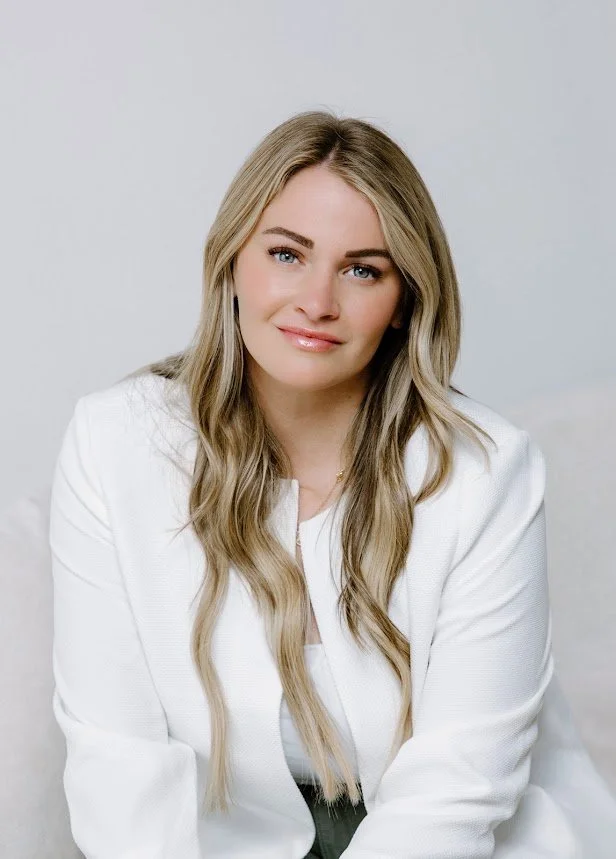 A young woman with long blonde hair, blue eyes, and fair skin posing against a light gray background. She is wearing a white blazer and has a slight smile.