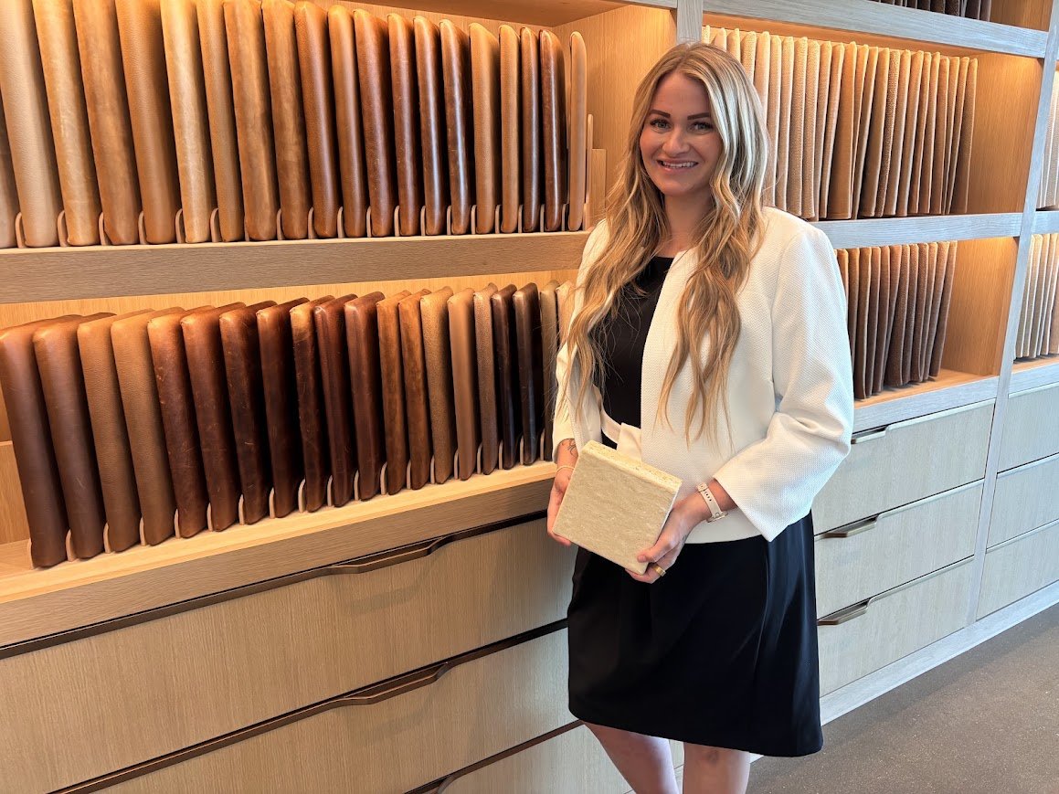 A woman with long blonde hair, wearing a white blazer and black dress, holding a beige notebook, standing in front of a display of various leather or fabric swatches arranged on shelves.
