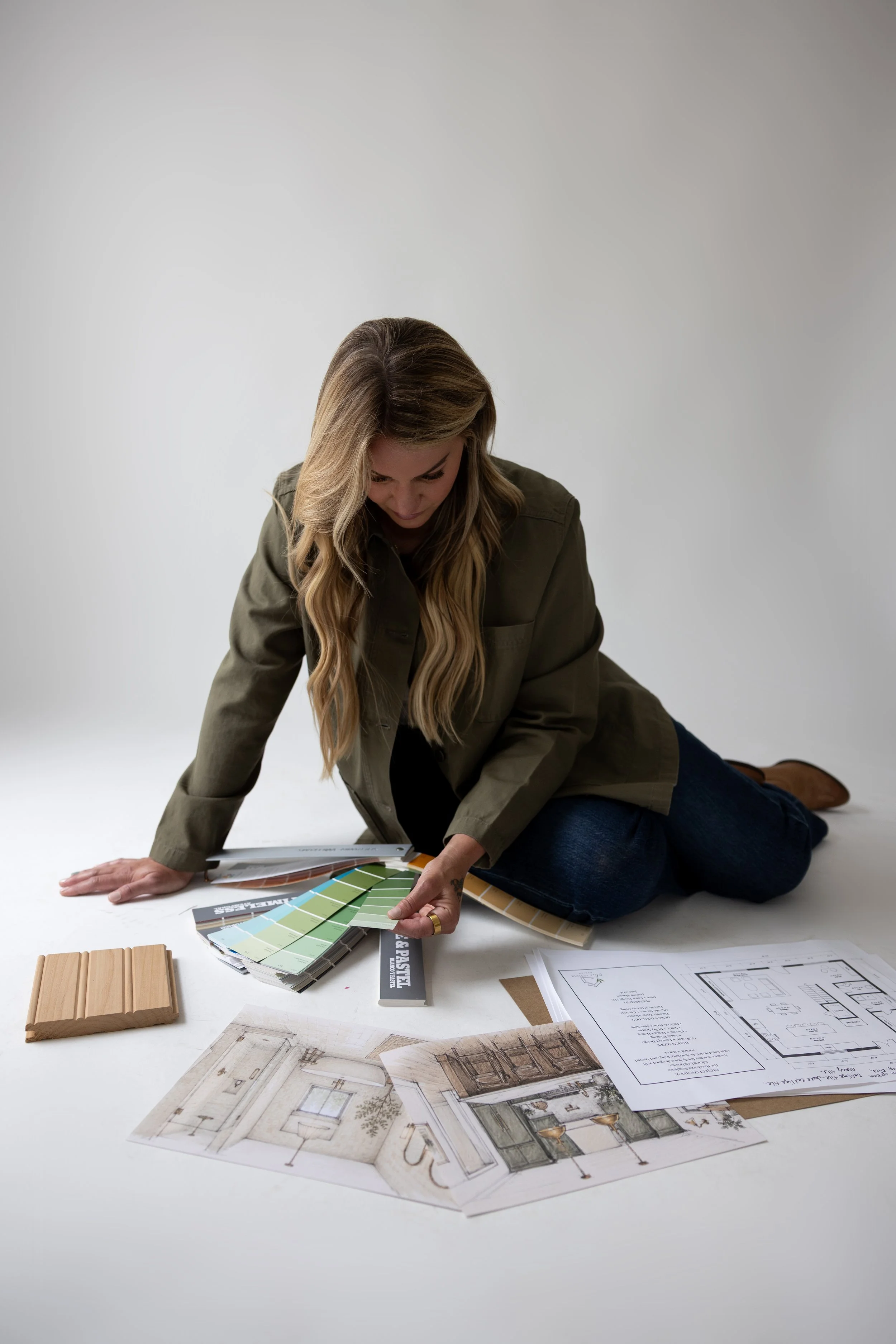 A woman in a casual jacket and jeans sitting on the floor, holding color swatches and architectural plans, surrounded by design and construction materials.