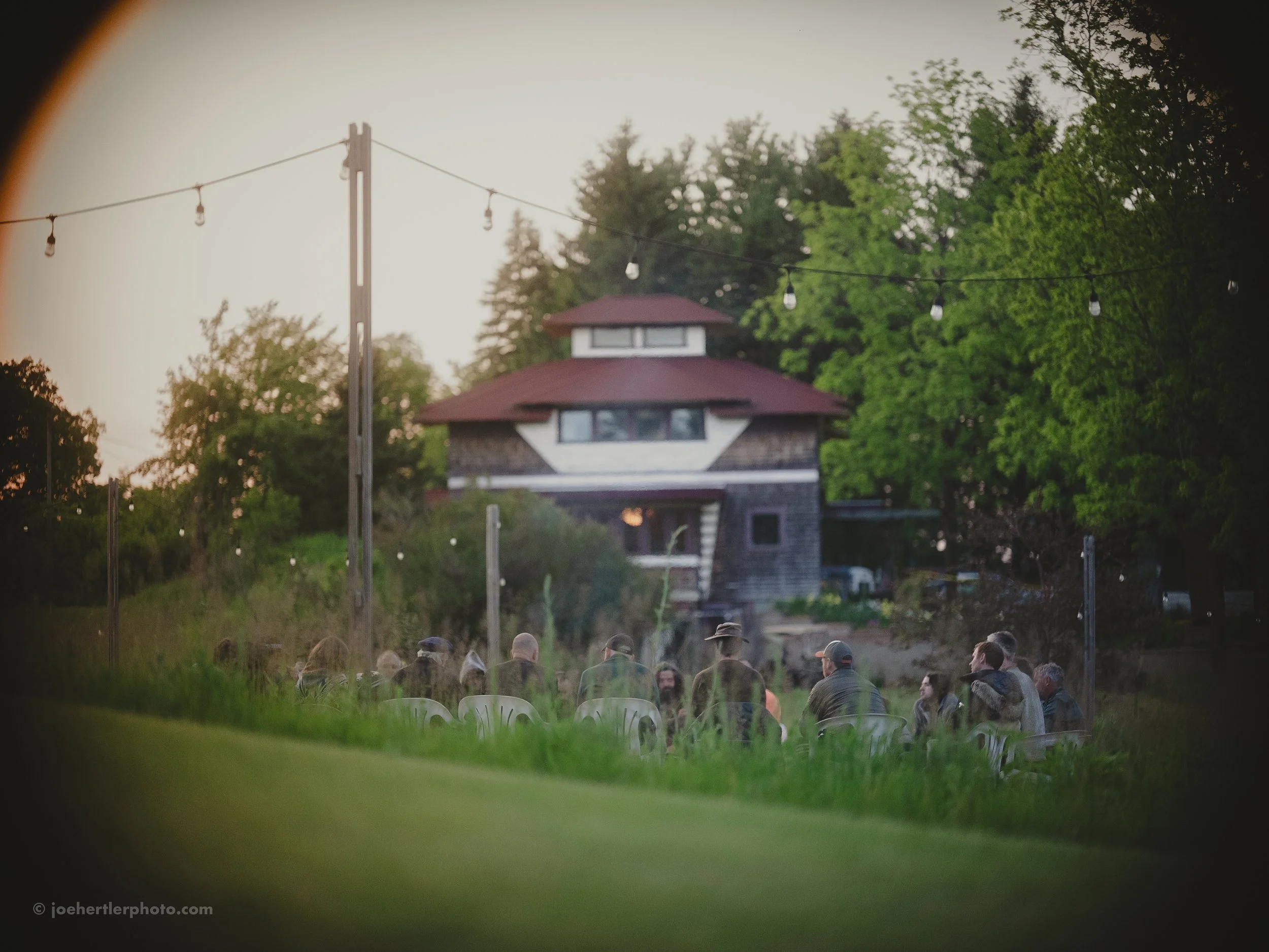 People seated outdoors in front of a house, seen through a circular or telescope view, with trees and string lights overhead.