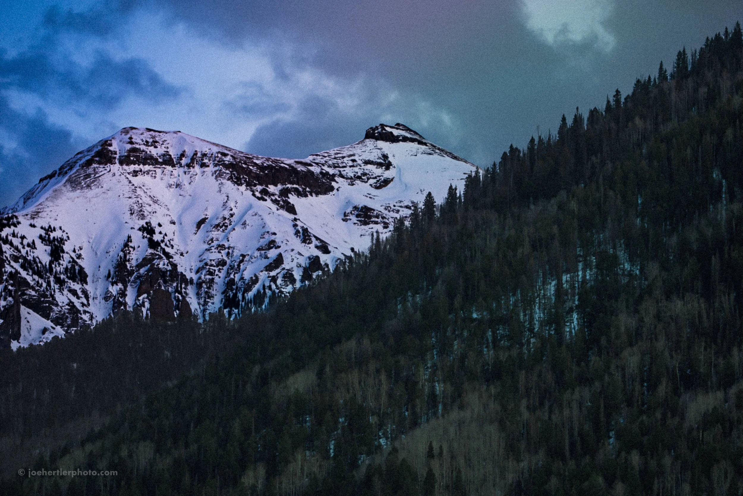 Snow-capped mountain peaks with dark forested slopes in foreground under cloudy sky.