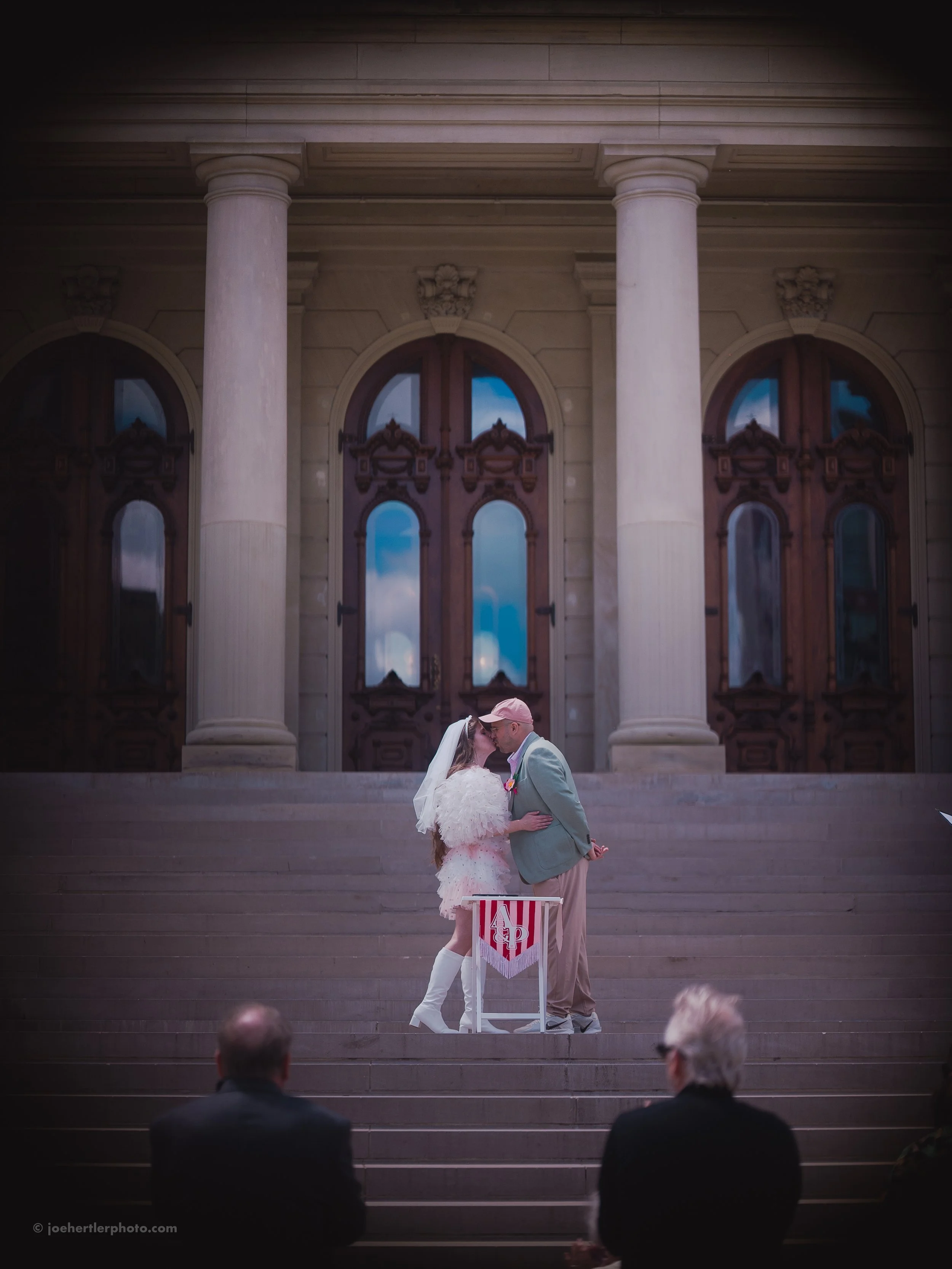 A bride and groom share a kiss during their wedding ceremony on the steps in front of a large historic building with columns and arched windows.