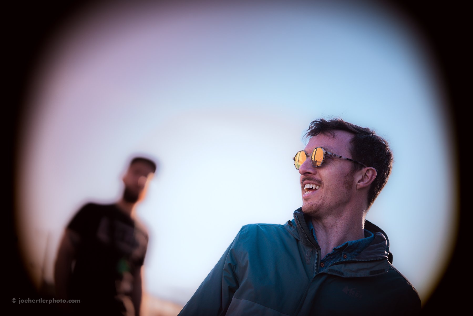 Two young men smiling and talking outdoors, one wearing reflective sunglasses and a blue jacket, the other slightly blurred in the background, against a clear sky.