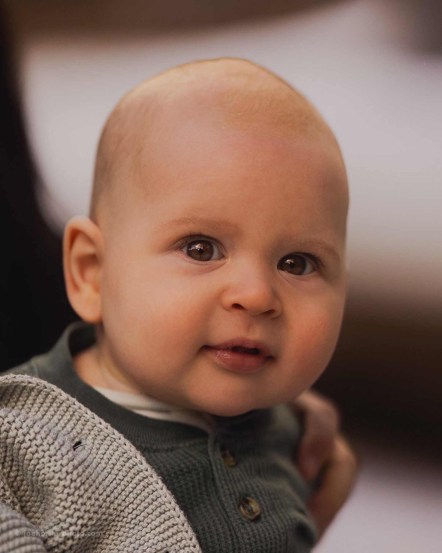Close-up of a baby with big brown eyes, light skin, and a bald head, wearing a dark gray shirt with buttons.