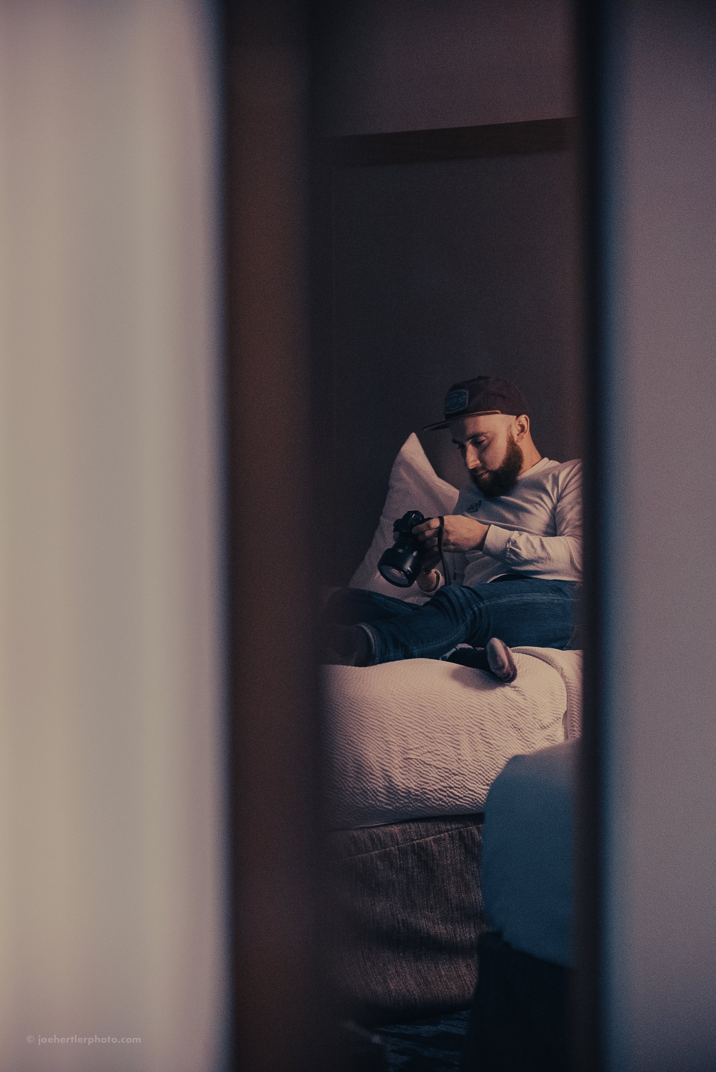 A man sitting on a bed, viewing a camera, seen through a door or closet door frame in a dimly lit room.