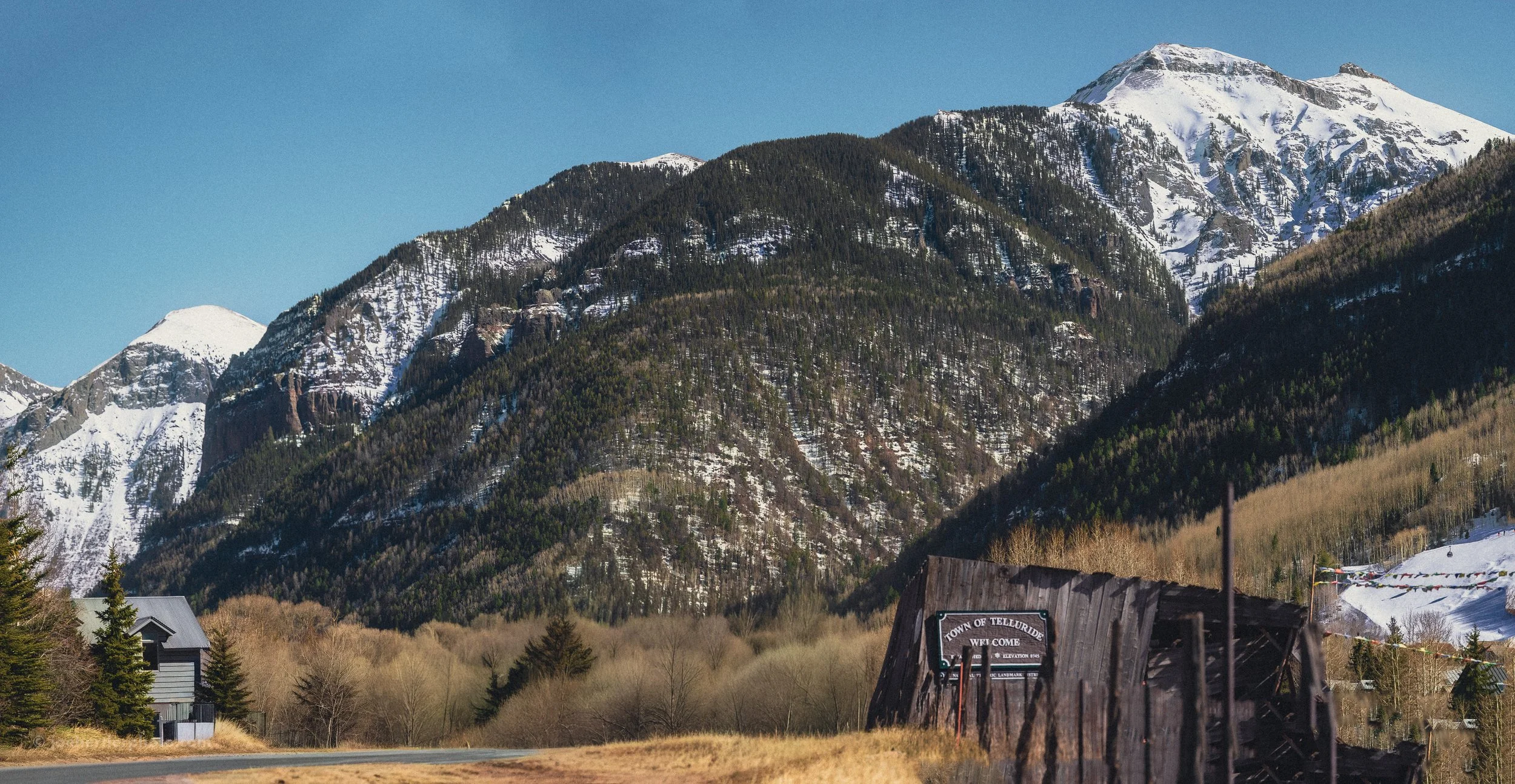Mountain landscape with snow-capped peaks, forested slopes, and a rural small town with houses and a weathered building in the foreground, under a clear blue sky.