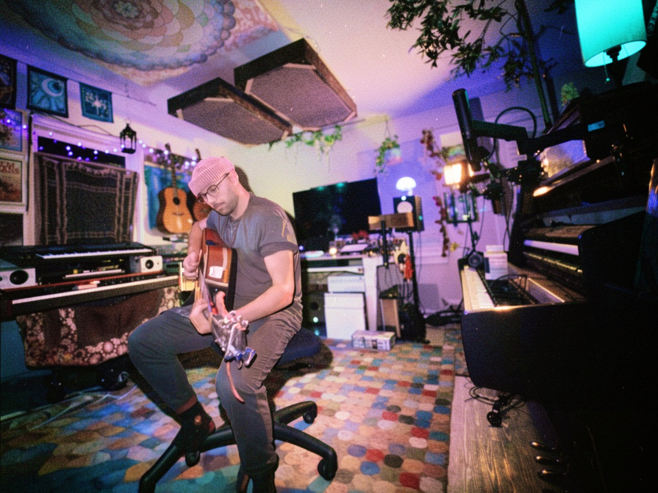 A man wearing glasses, a pink beanie, and casual clothes is sitting on an office chair in a music studio, playing an acoustic guitar. The studio has multiple musical instruments, including pianos and guitars, and is decorated with guitars on the wall, colorful lights, plants, and framed artwork.