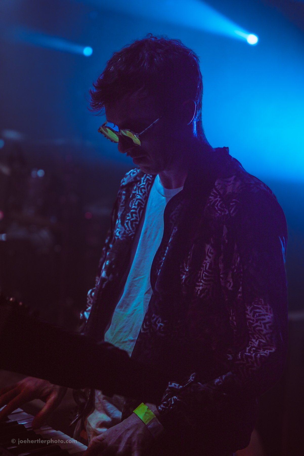 A dark scene with a person wearing sunglasses, playing a keyboard under blue stage lighting.