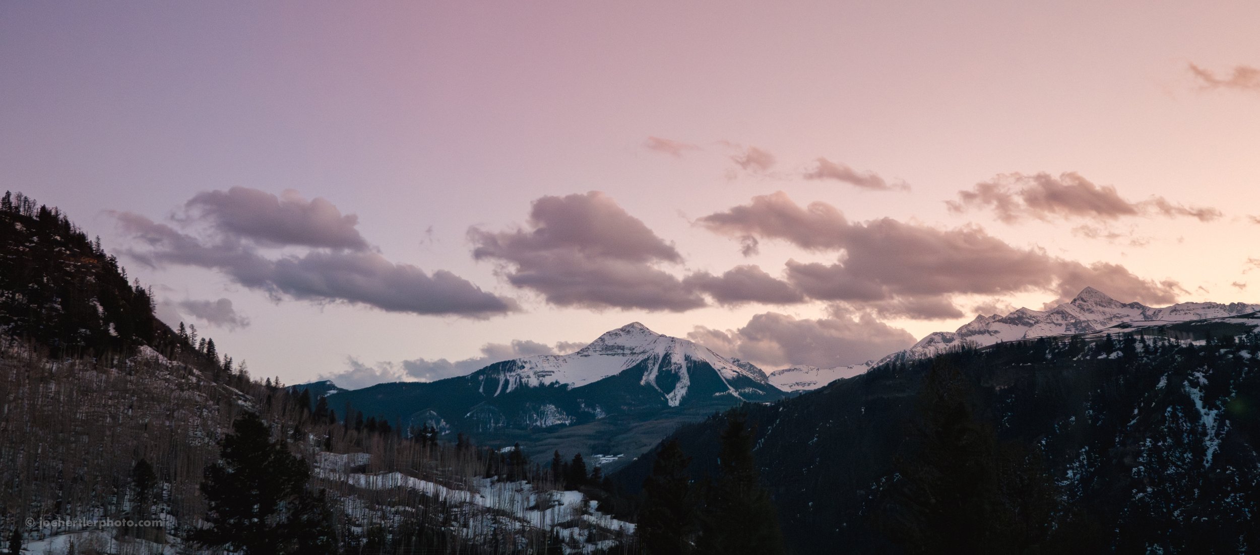 Snow-capped mountains under a pink and purple sky with scattered clouds, forested slopes in the foreground.