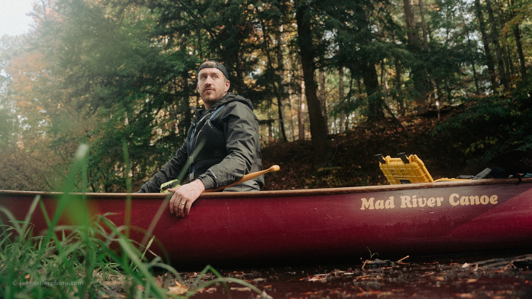 A man sitting in a red canoe on a river, surrounded by trees with fall foliage.