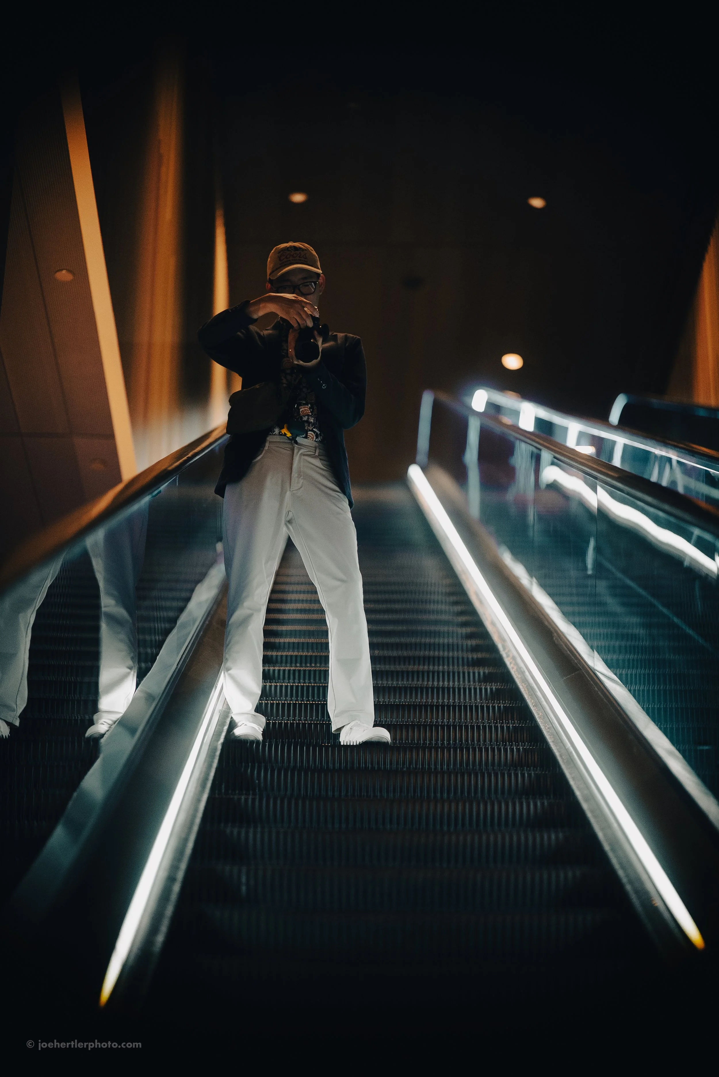 Person wearing a beige cap and black jacket taking a photo on an escalator inside a modern building.