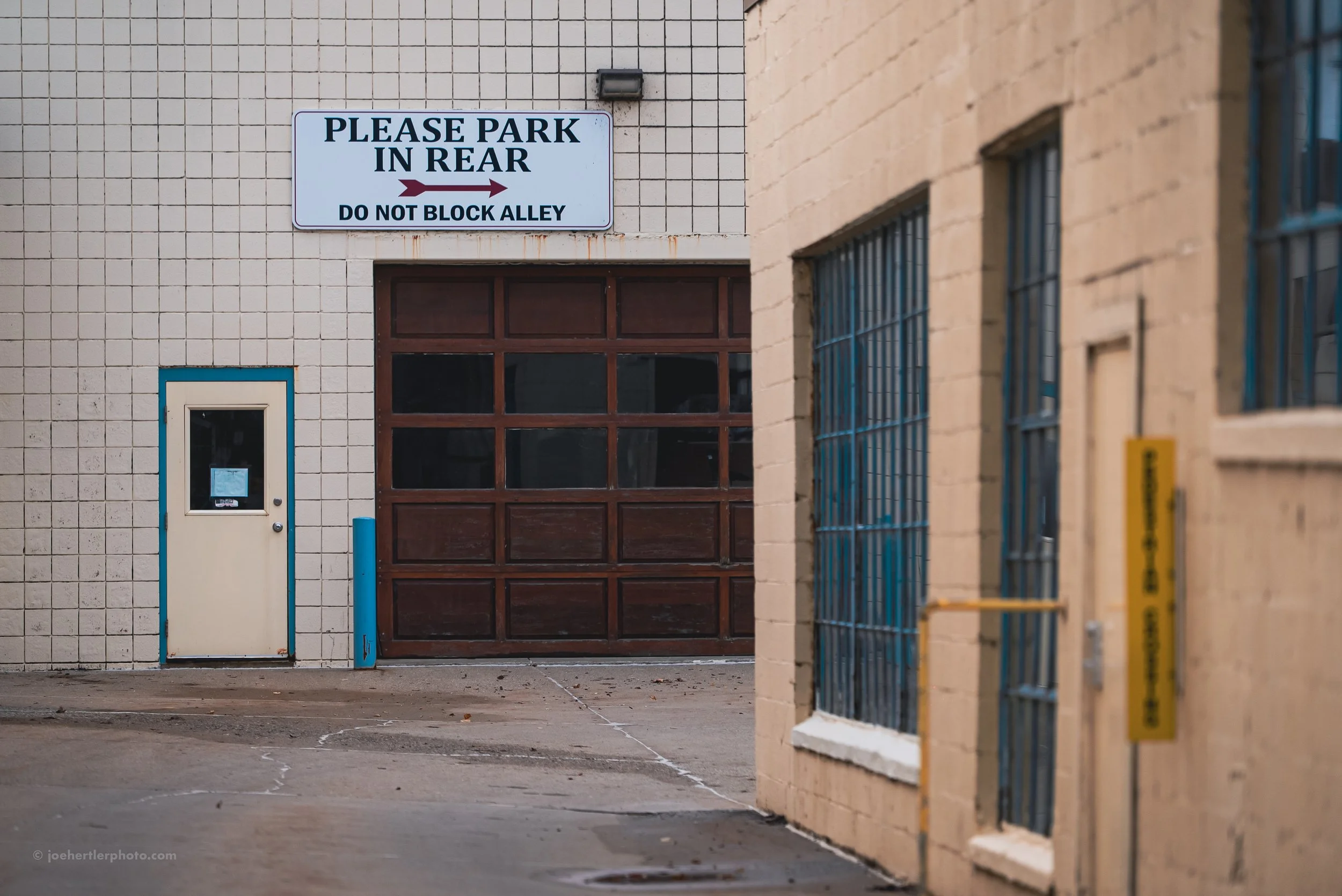 A loading dock area with a sign saying "Please park in rear, do not block alley." There is a beige door with a small window on the left, a large brown garage door in the center, and windows with security bars on the right.