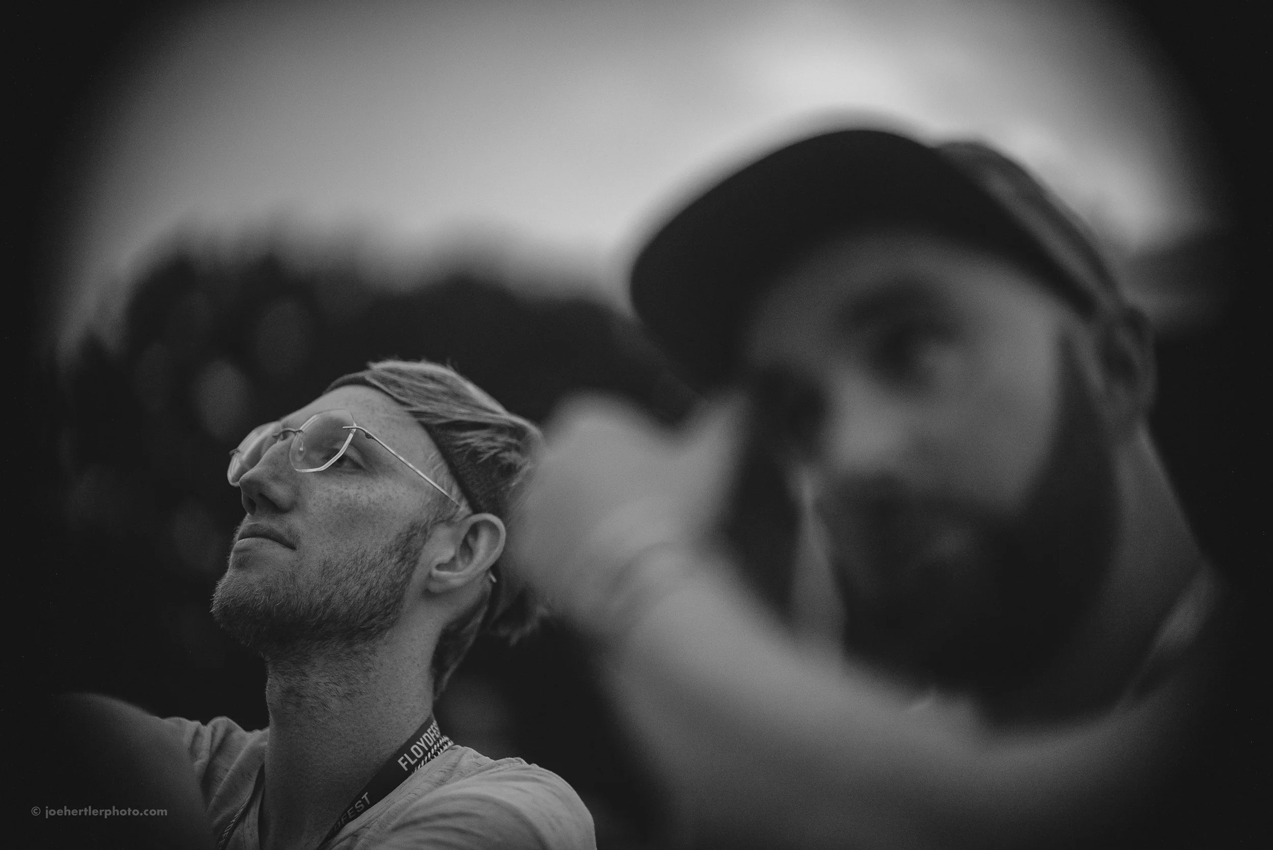 Black and white photo of two young men, one in the foreground with a cap, beard, and glasses, slightly smiling with closed eyes, and another in the background with a hat, looking directly at the camera, with a blurred outdoor scenery.