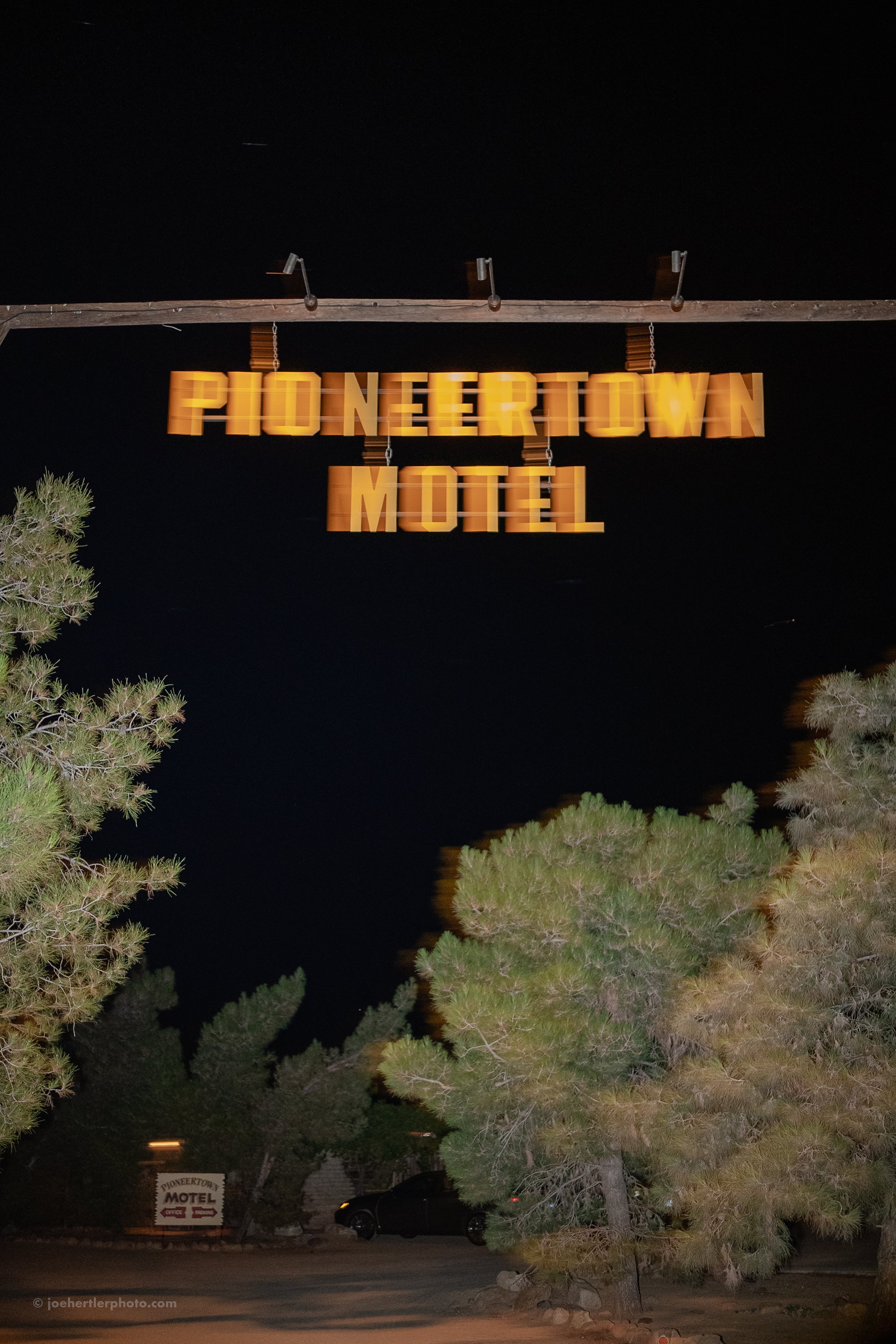 Nighttime exterior view of the Pioneertown Motel sign hanging above trees and parked cars.