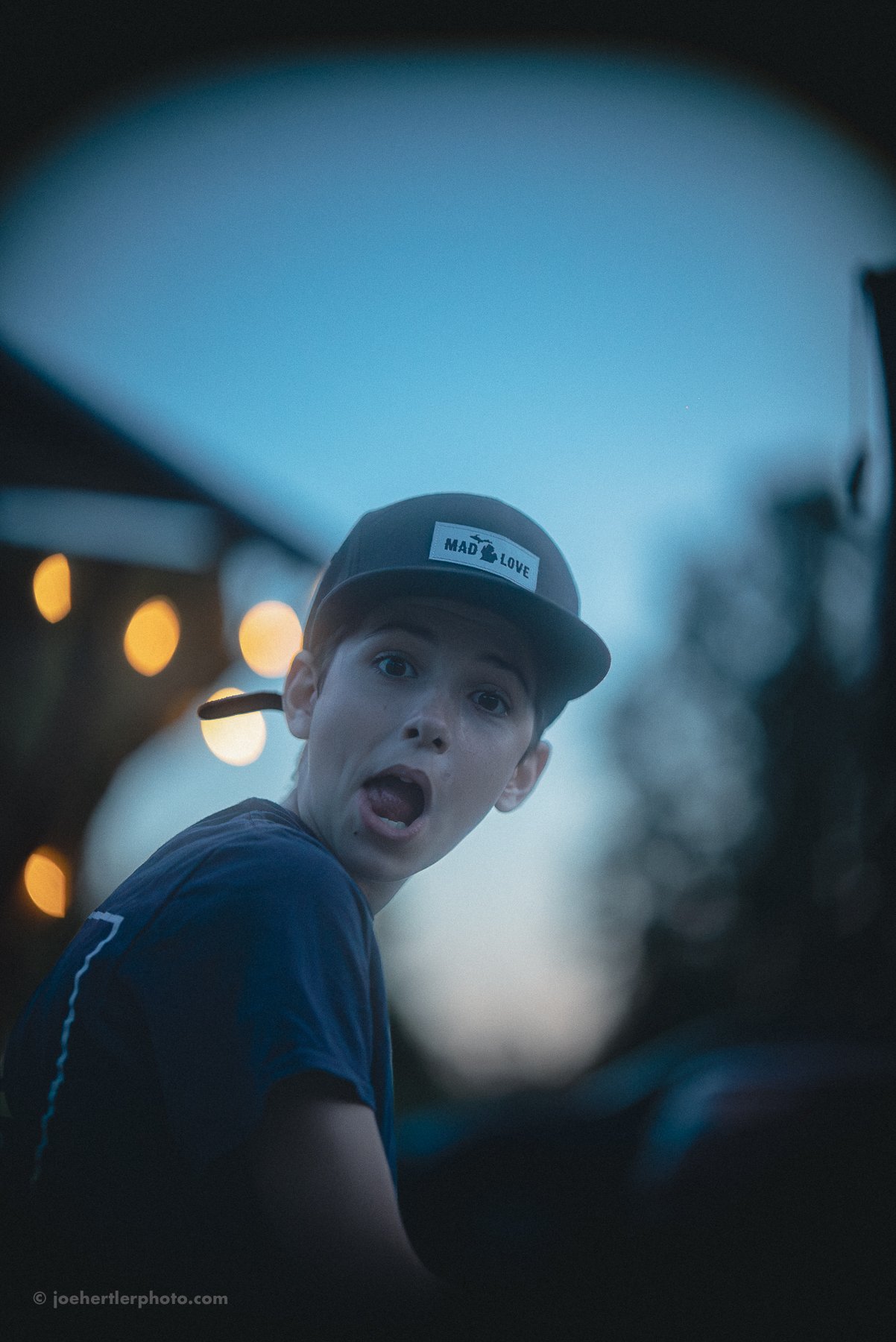 A young man wearing a black cap with the words "MAD LOVE" on it, making a surprised or animated facial expression, with a blurred background of orange bokeh lights and a dusk sky.
