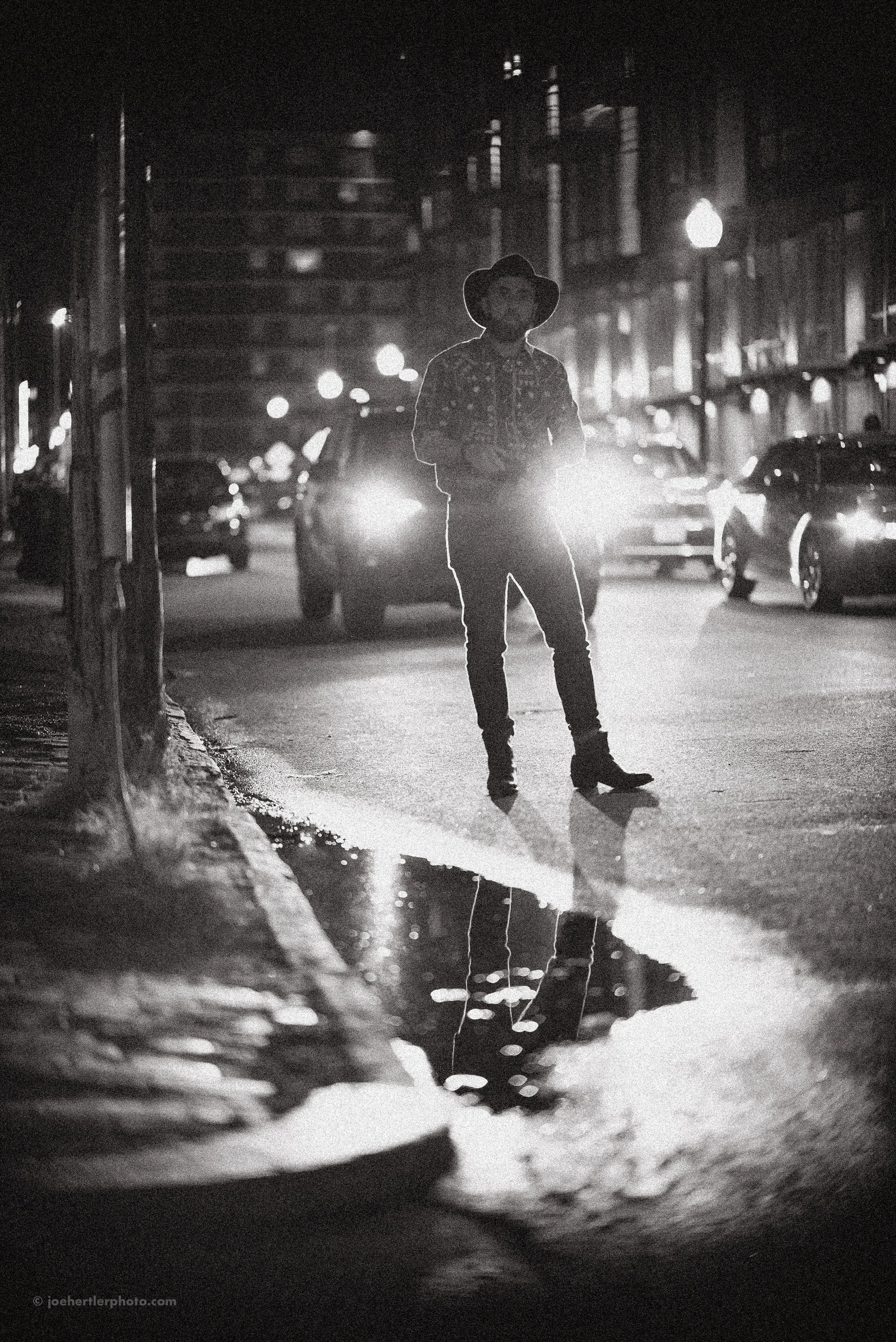 A person wearing a hat standing on a city street at night, backlit by car headlights, with their reflection visible in a puddle on the ground.