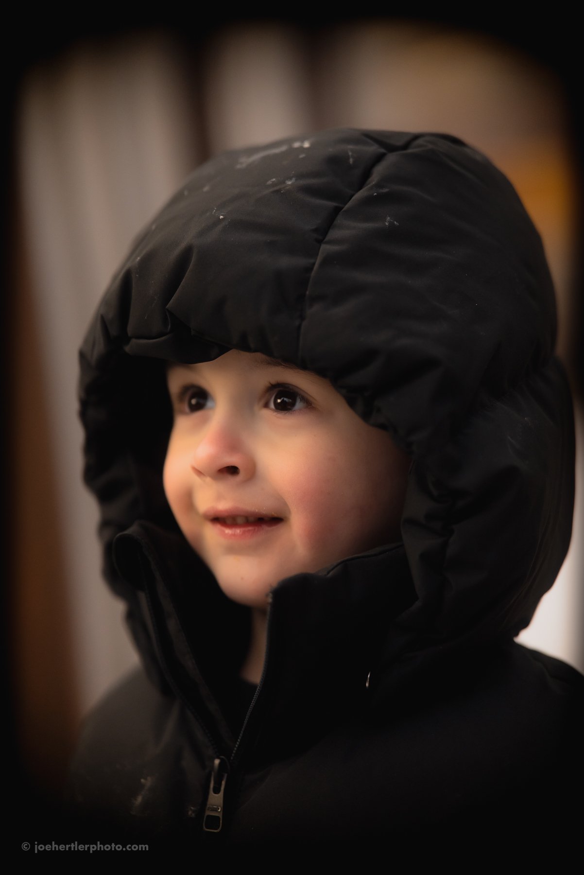 Young child smiling and looking up, wearing a black winter coat with a hood over their head.