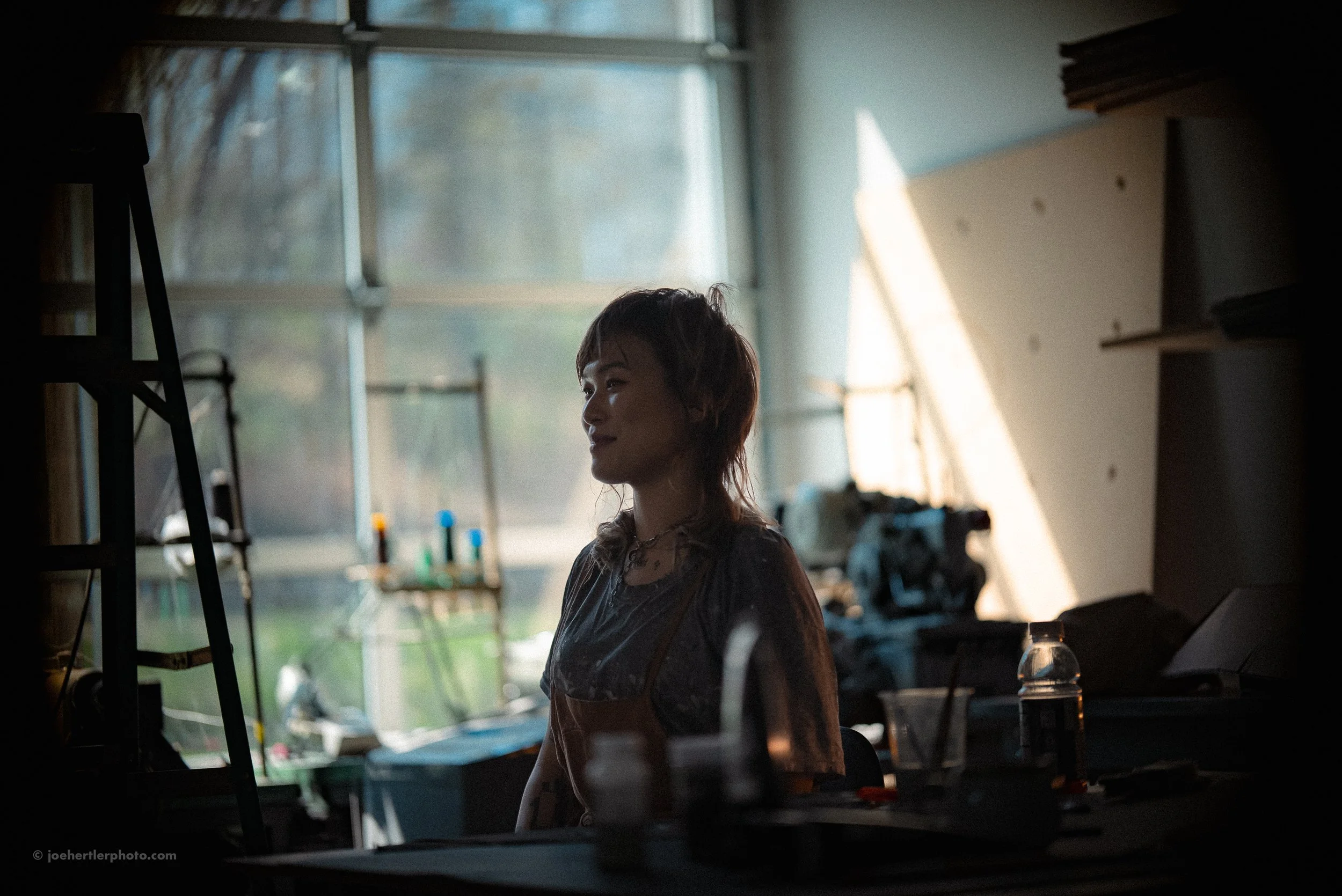 A woman with short, wavy hair stands in a dimly lit room with large windows, with sunlight coming in from the side. The room contains various objects and equipment on shelves and tables, suggesting a workshop or studio setting.