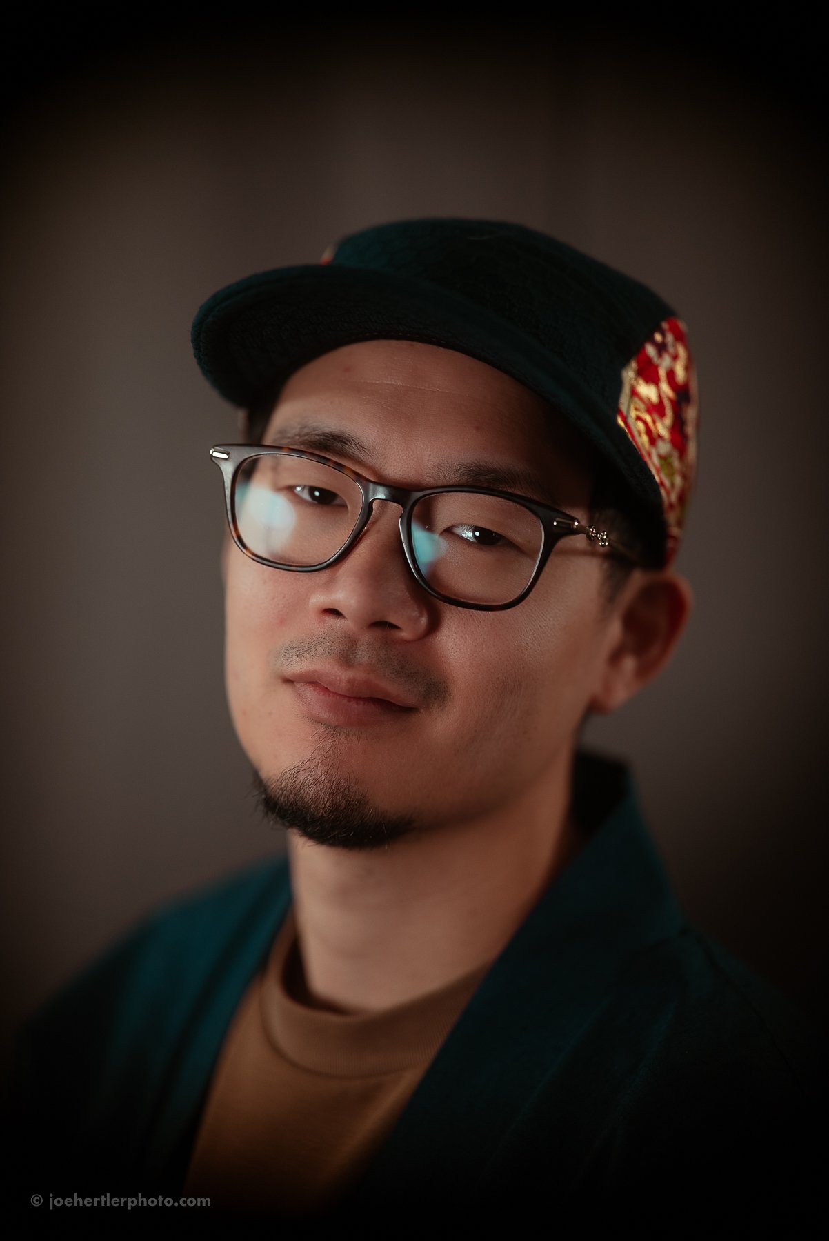 Portrait of a man wearing glasses, a black cap with a red and gold design on the side, and a dark jacket, against a dark background.