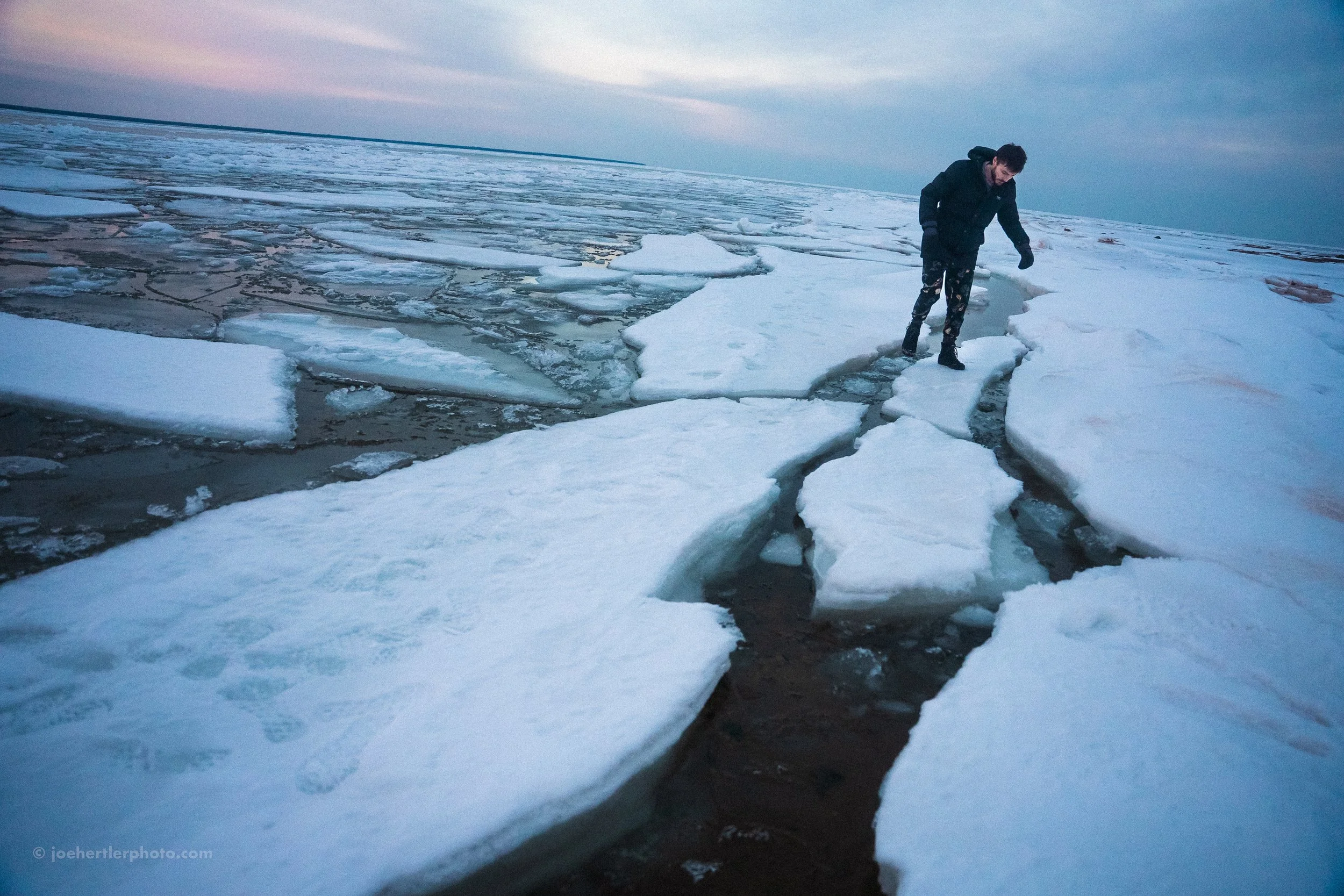 A person walking on large, broken ice floes in a cold, open body of water during twilight.
