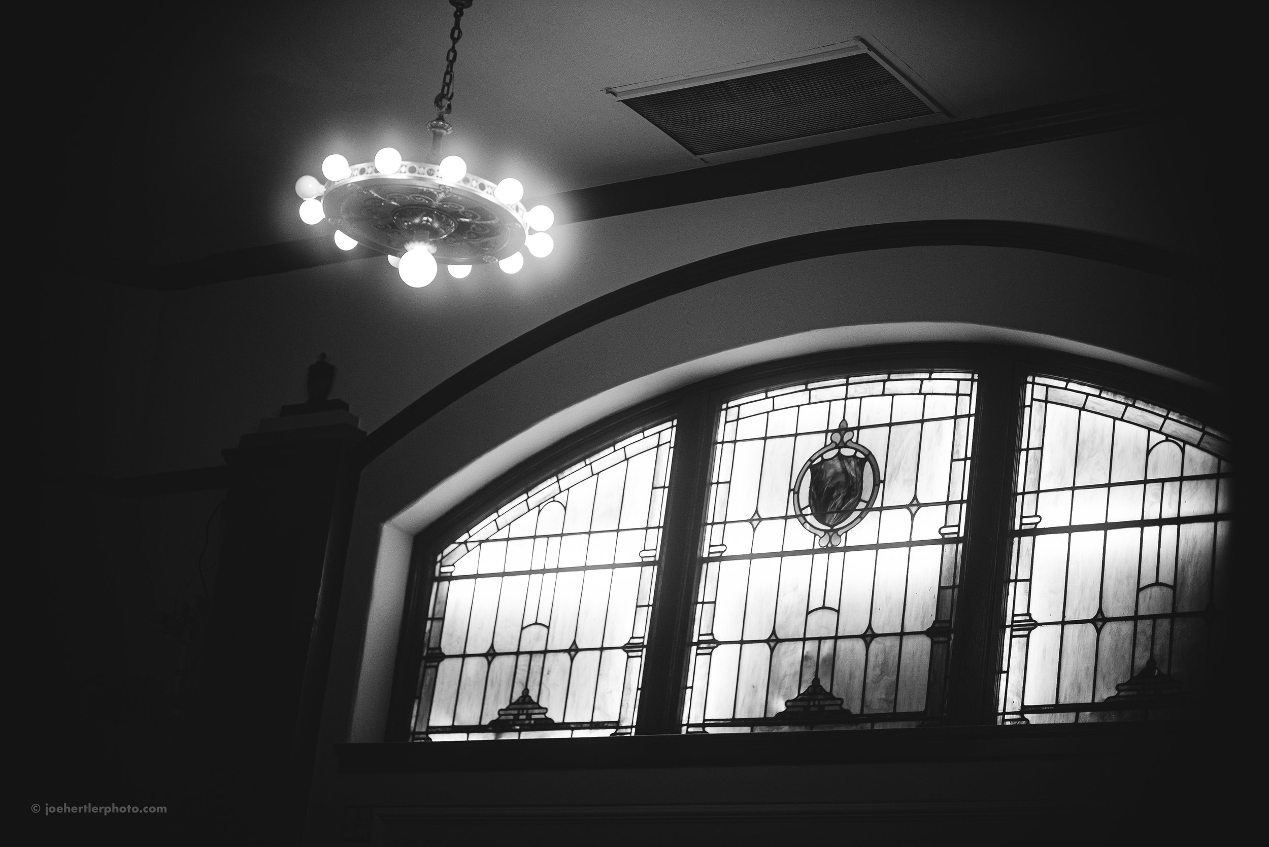Black and white photo of an interior ceiling with a chandelier hanging and a stained glass window below.