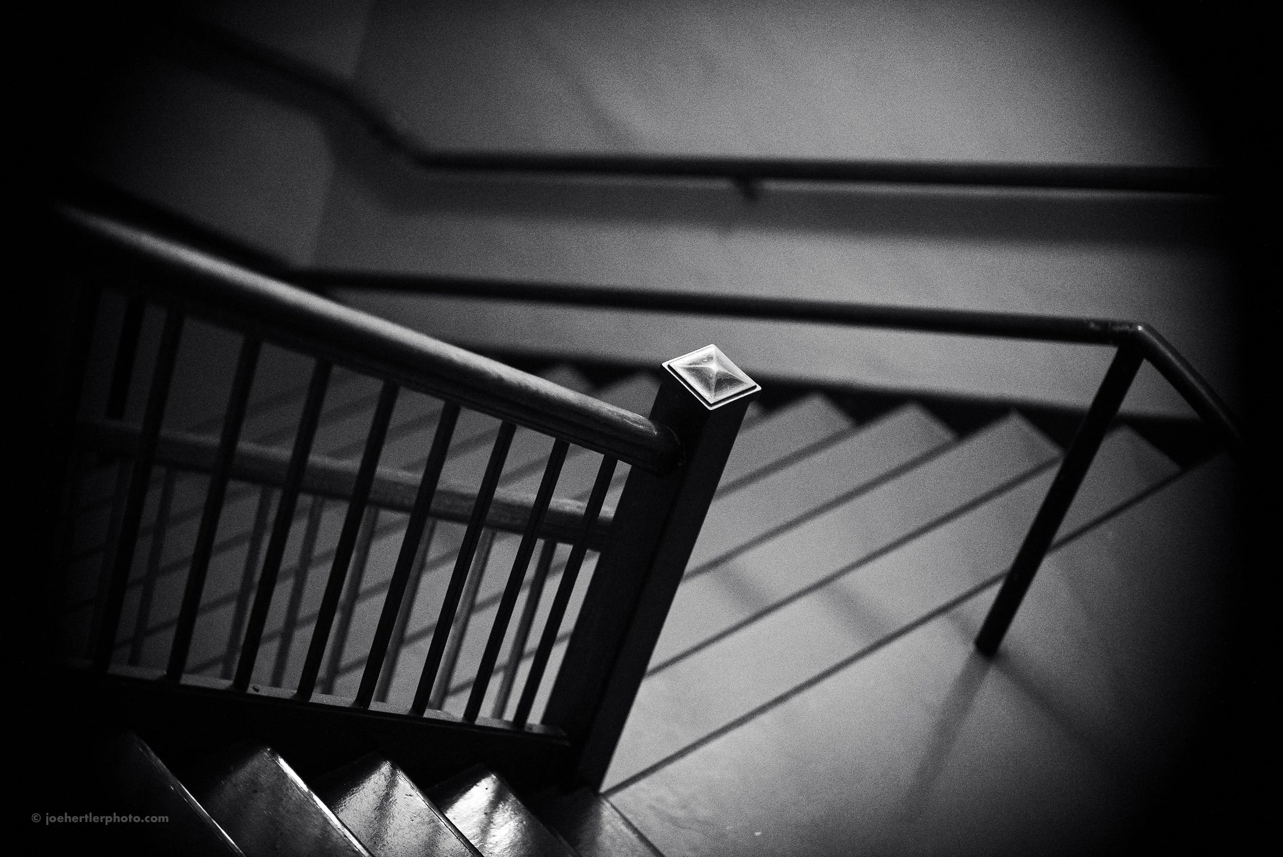 Black and white photo of an empty staircase viewed from above, with shadows cast by the railing.