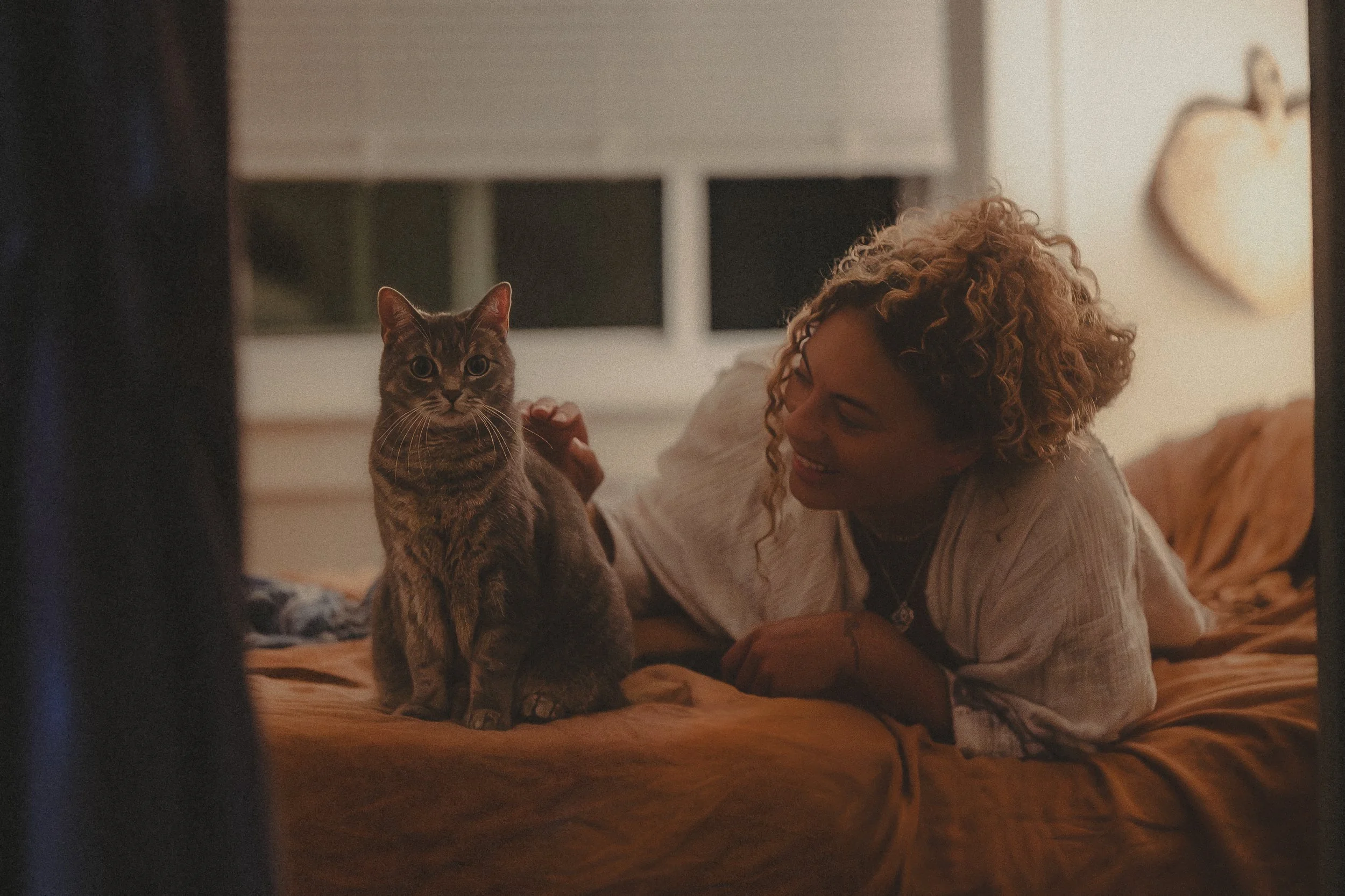 A woman with curly blonde hair lying on a bed, smiling at a gray tabby cat sitting on the bed next to her.