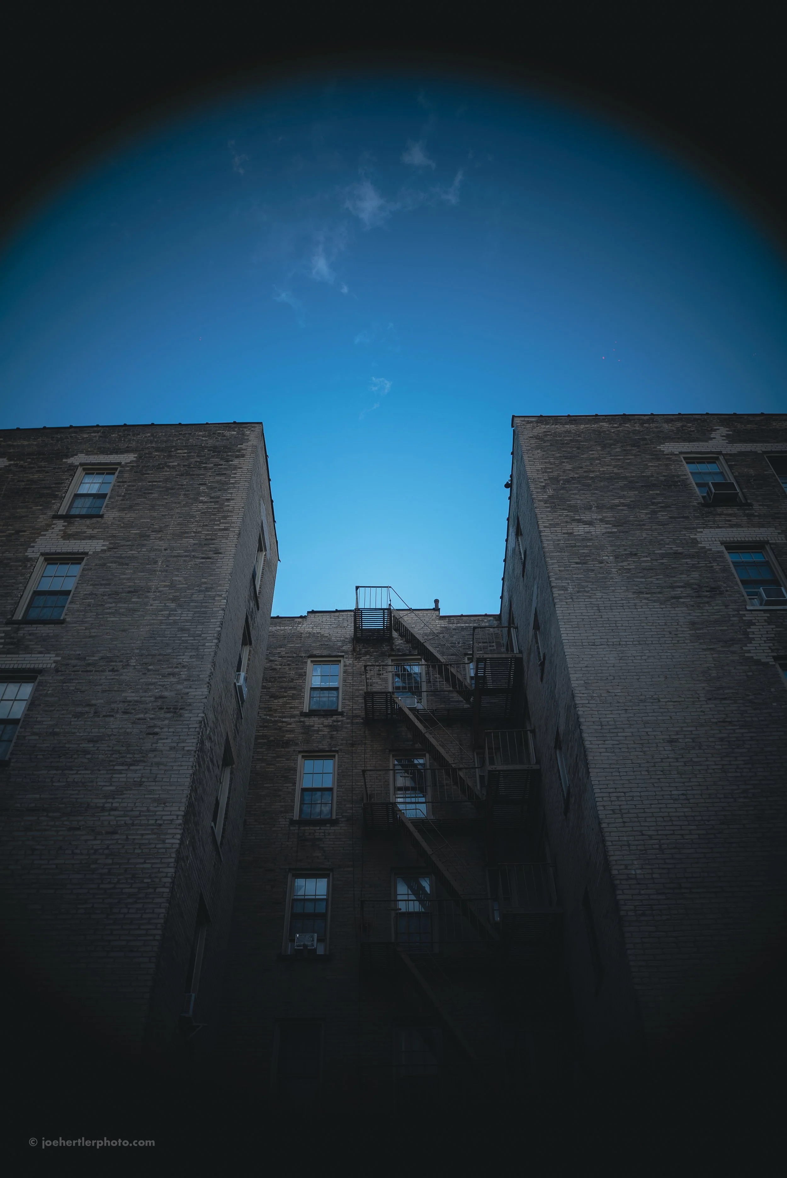 Looking up at an outdoor brick apartment building with fire escape stairs and windows, with a clear blue sky and a few clouds overhead.