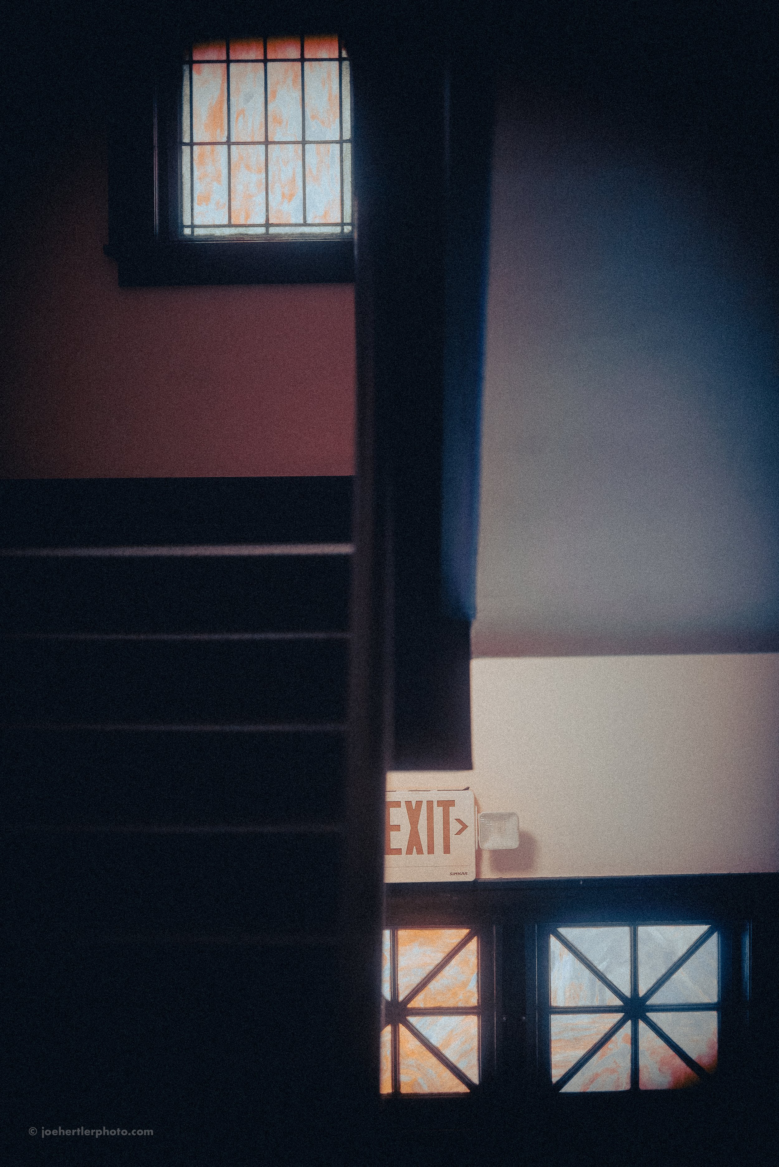 An interior view of a staircase with a small window at the top and a larger window at the bottom, with sunset or sunrise light visible through the windows, and an exit sign on the wall in between.