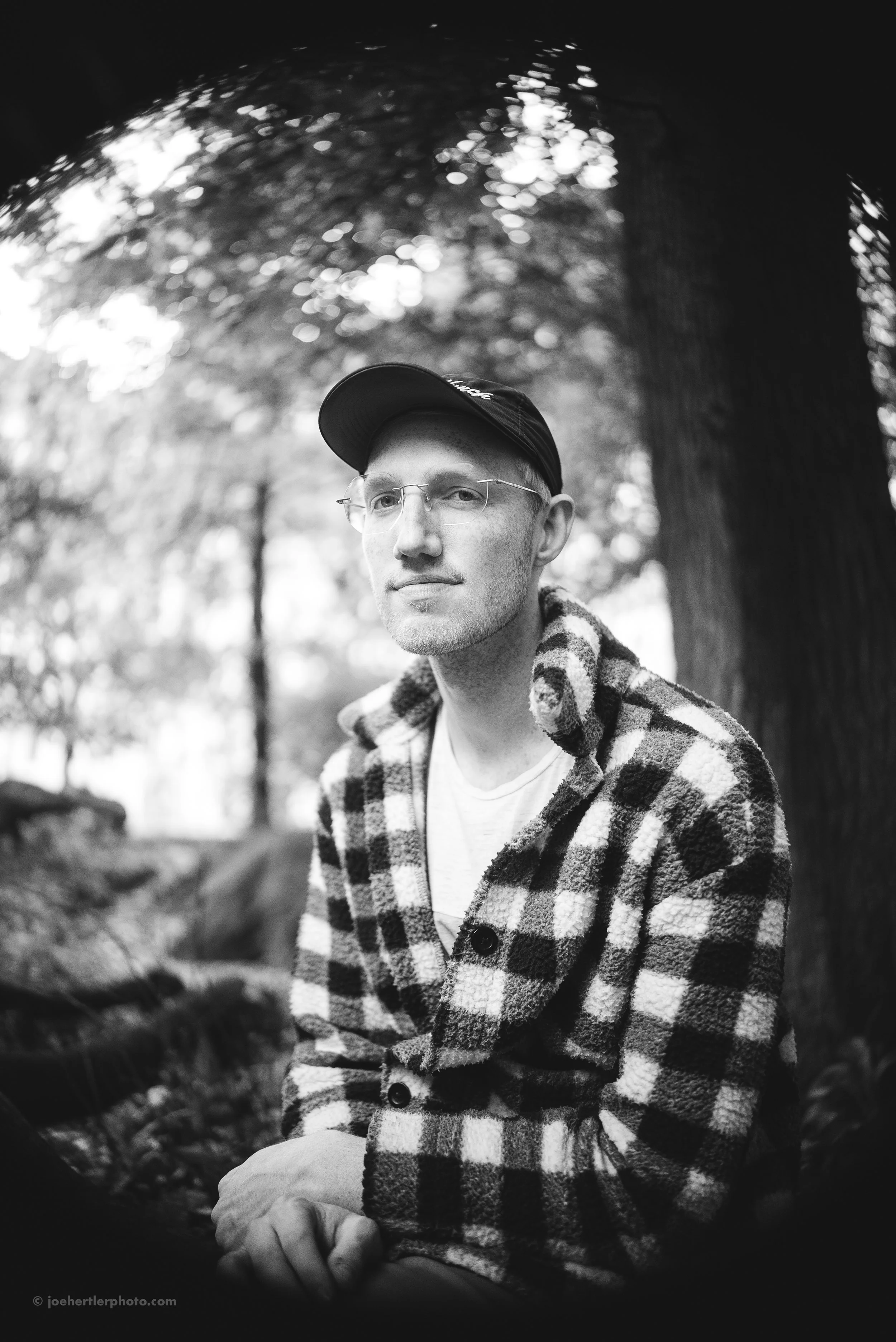 A black and white portrait of a man wearing glasses, a baseball cap, and a checkered flannel shirt, sitting outdoors with trees and blurred foliage in the background.