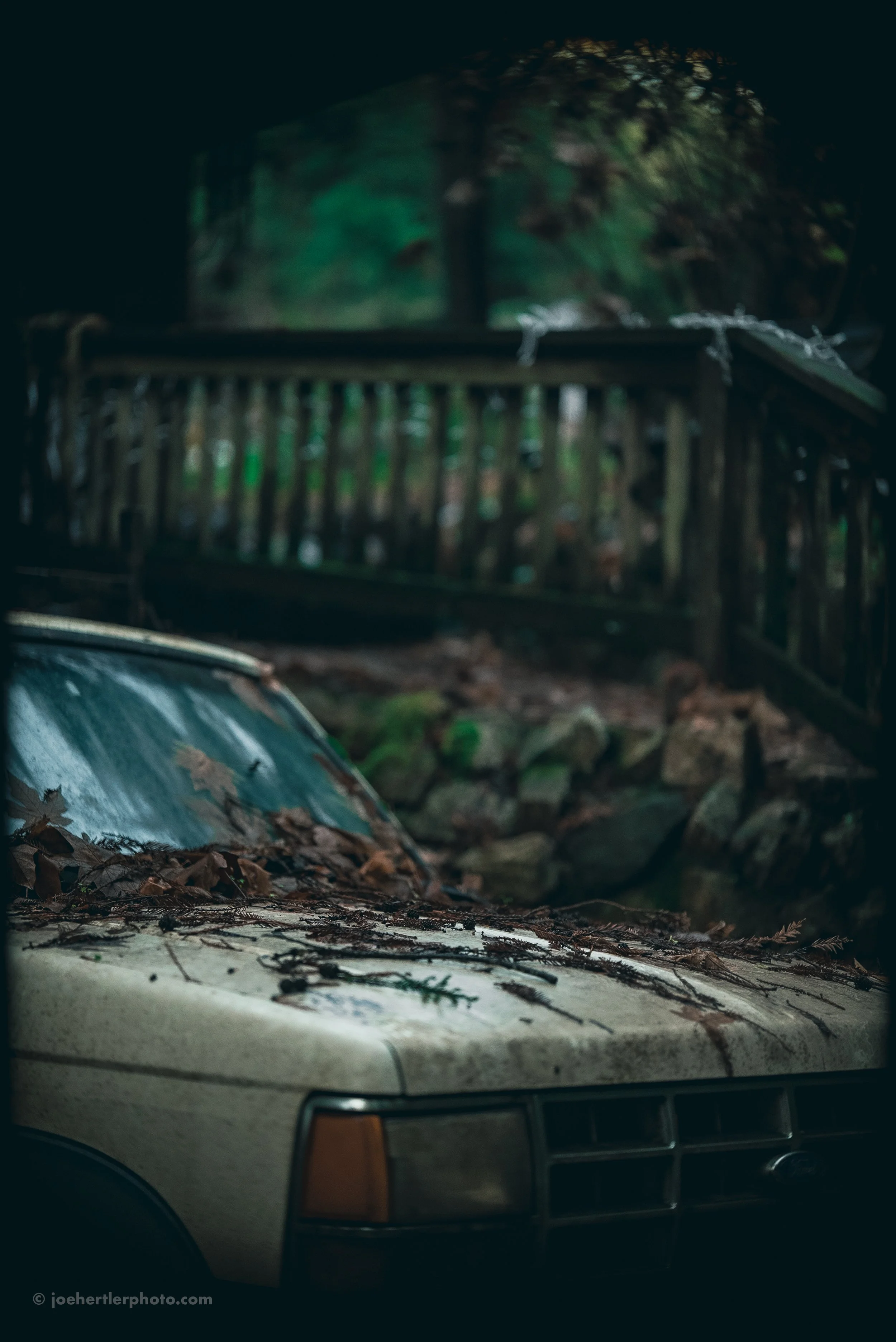 An old, dusty car covered with dry leaves and debris, parked outdoors near a wooden fence and rocks in a wooded area.