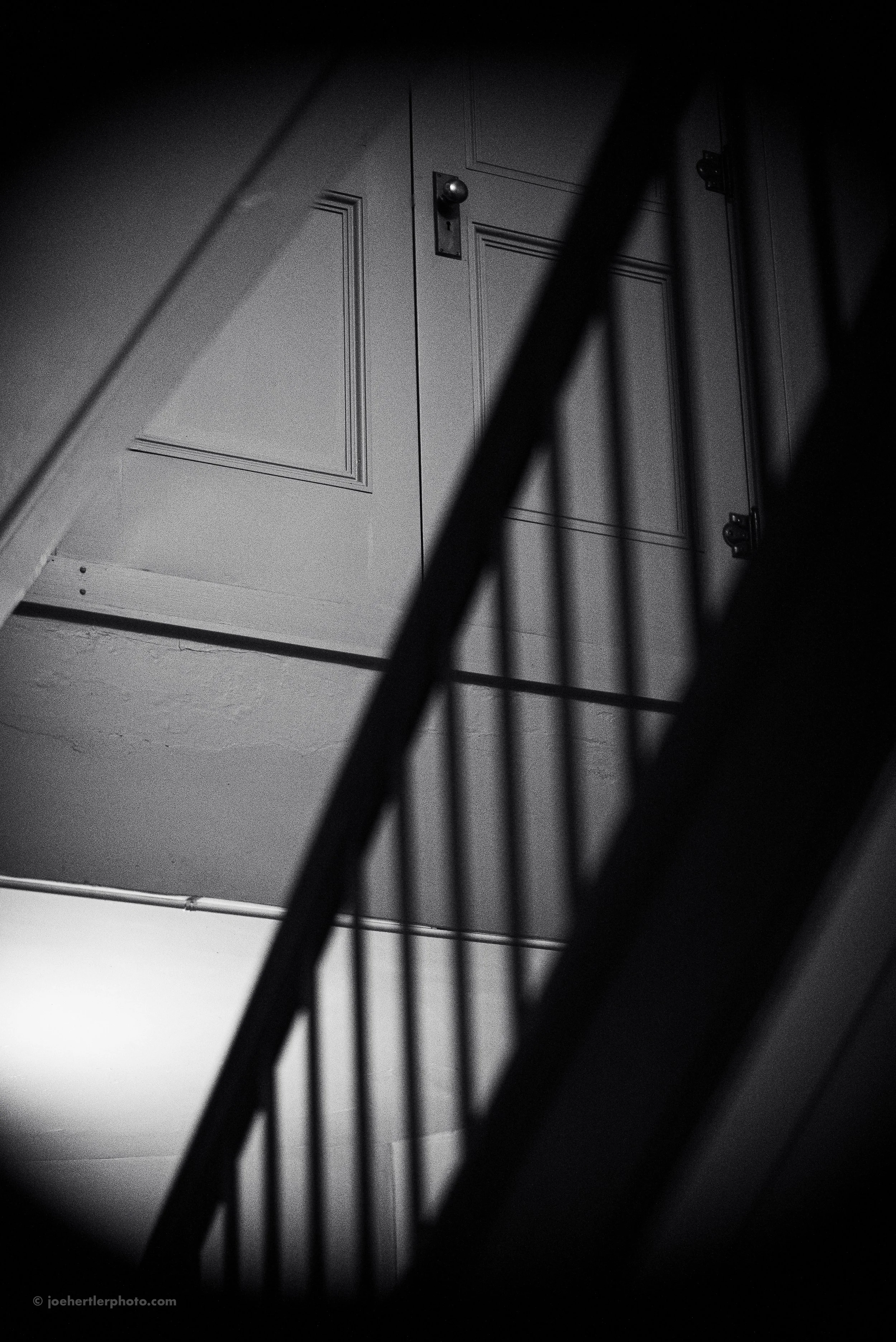 Black and white photo of a staircase with a railing, looking up towards a closed door with a round doorknob.