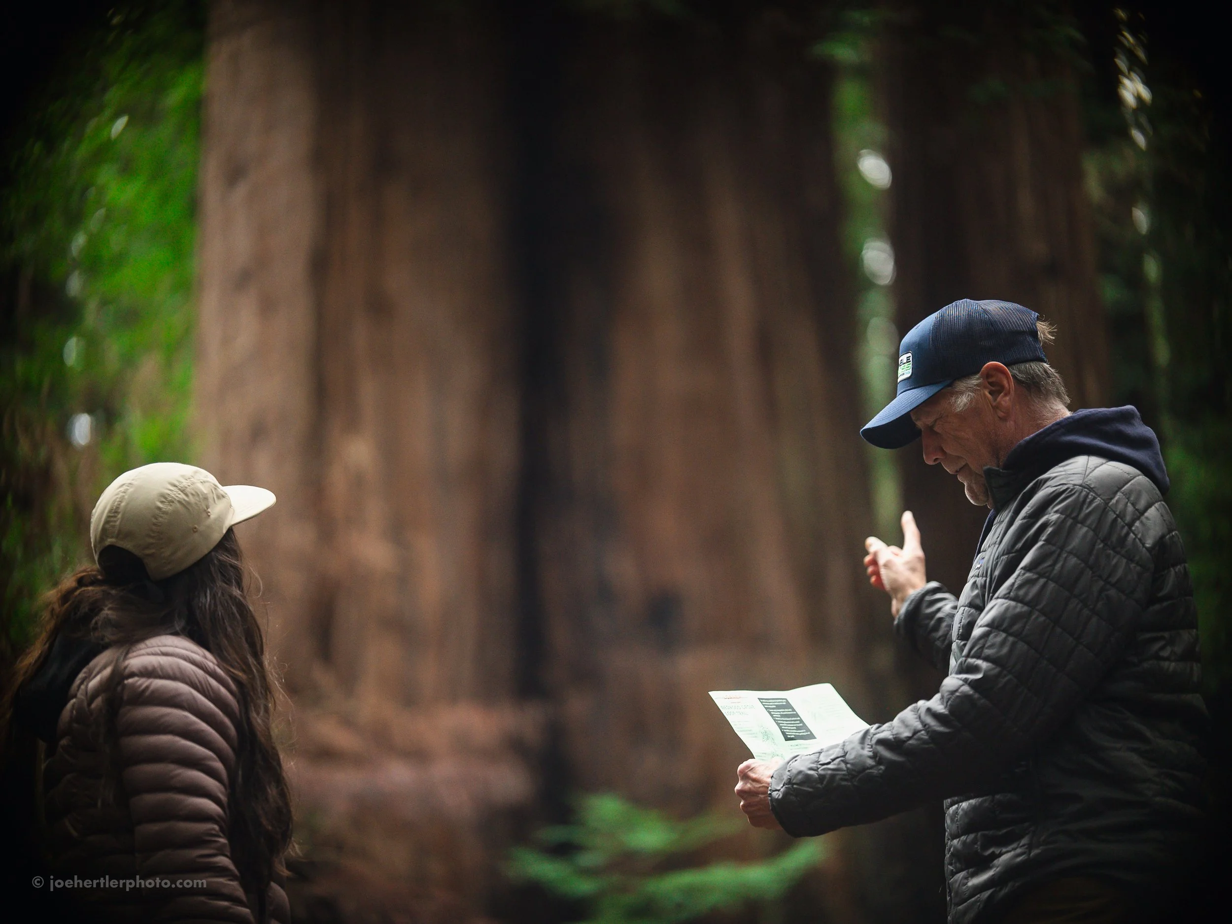 A man and a woman in outdoor clothing and hats, standing in a forest with large trees, engaged in a conversation or educational activity.