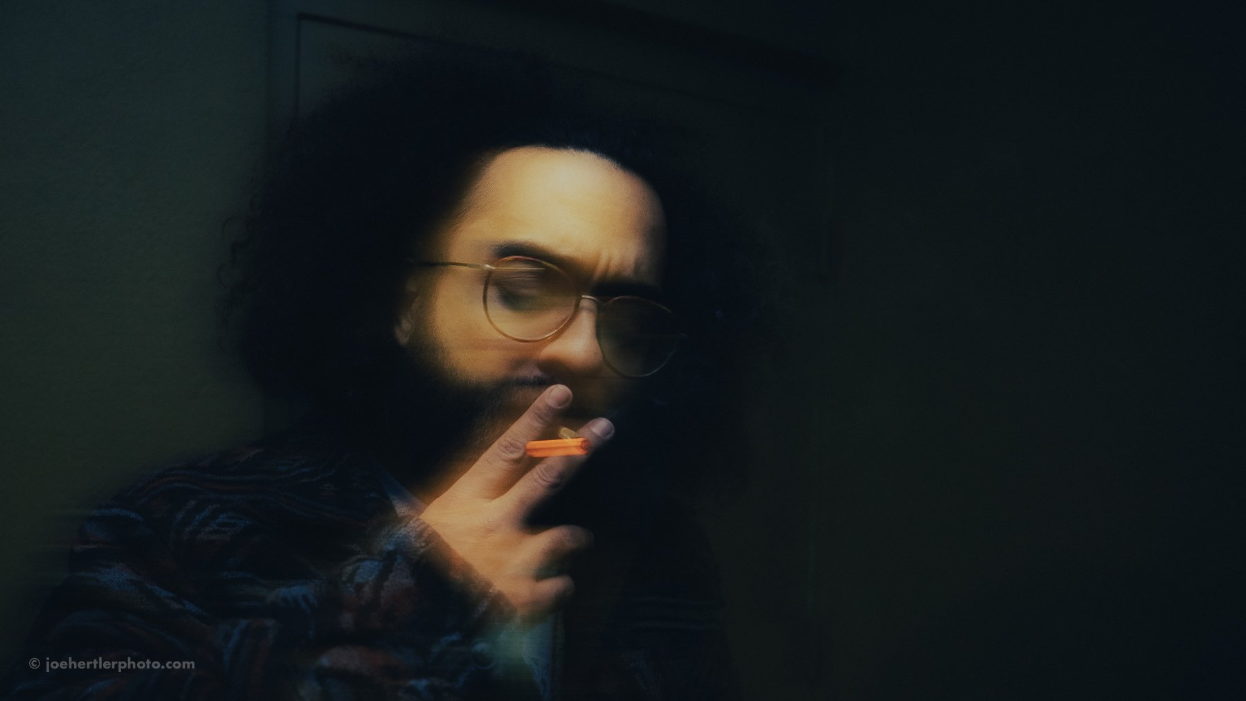 A young man with curly hair and glasses, smoking a cigarette in a dimly lit room.
