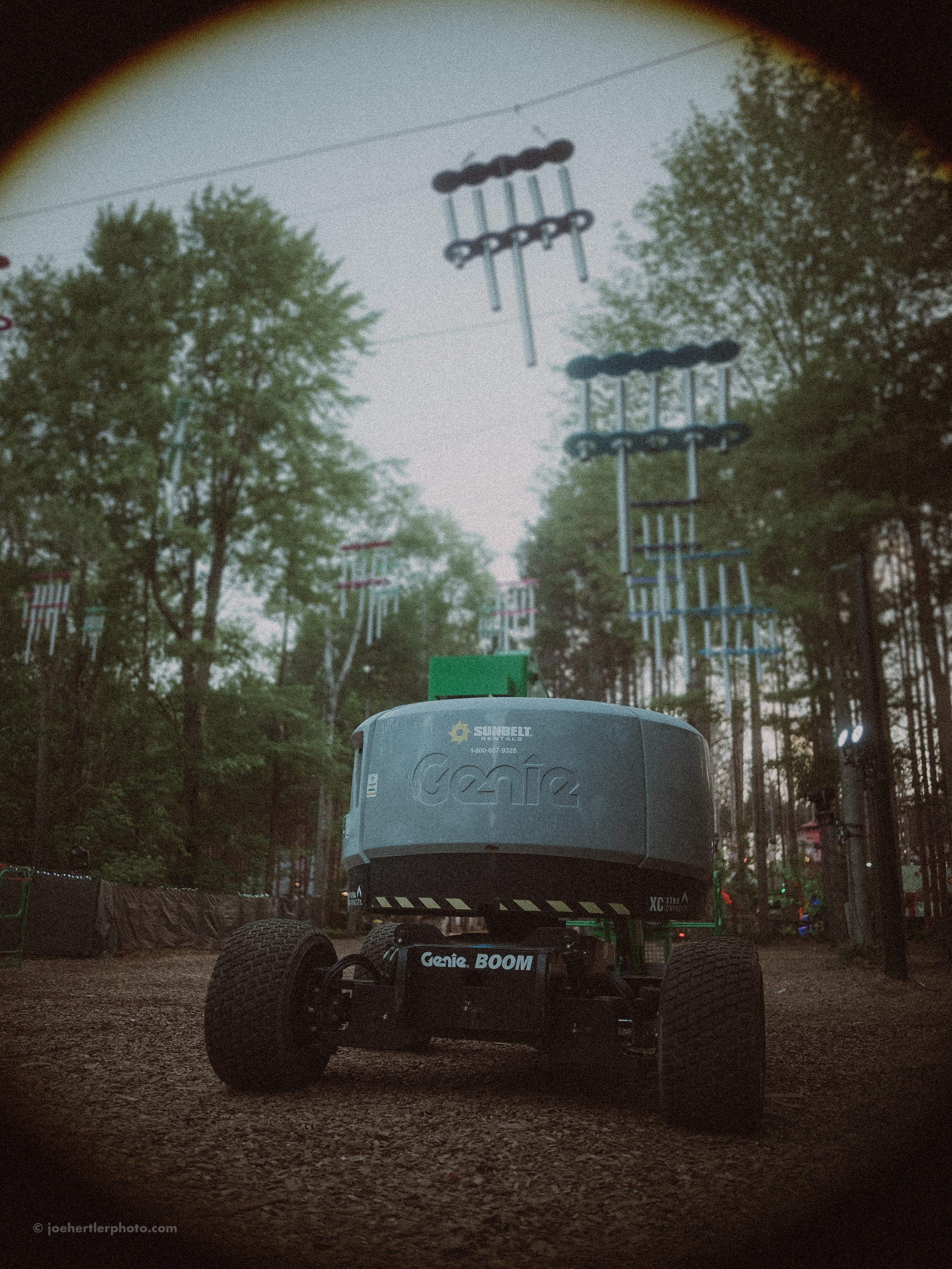An outdoor adventure park with a remote-controlled robot on a dirt ground, surrounded by tall trees, with zip lines and platforms in the background.
