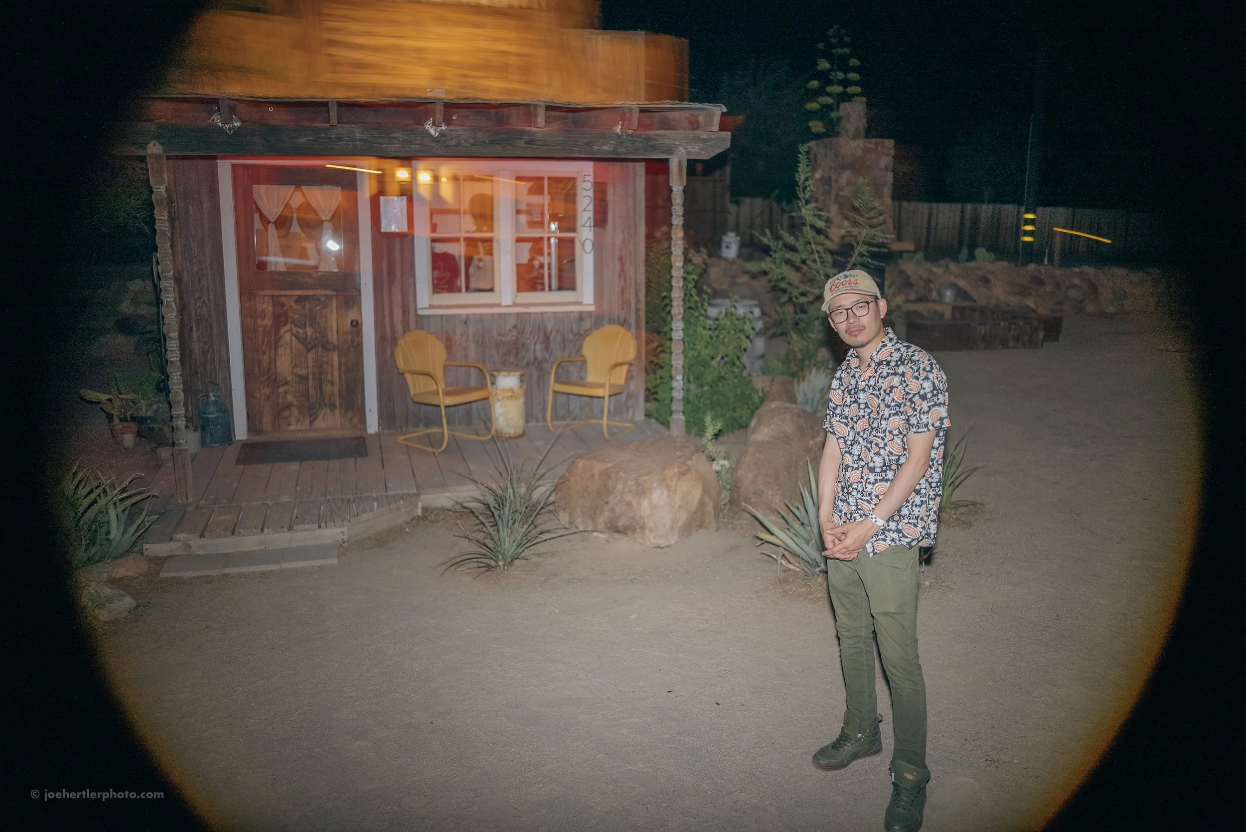 A man standing outside a small wooden house at night. The house has a porch with two yellow chairs and a small table between them, and a door with the number 5240. The man is wearing glasses, a patterned short-sleeve shirt, dark green pants, and a be